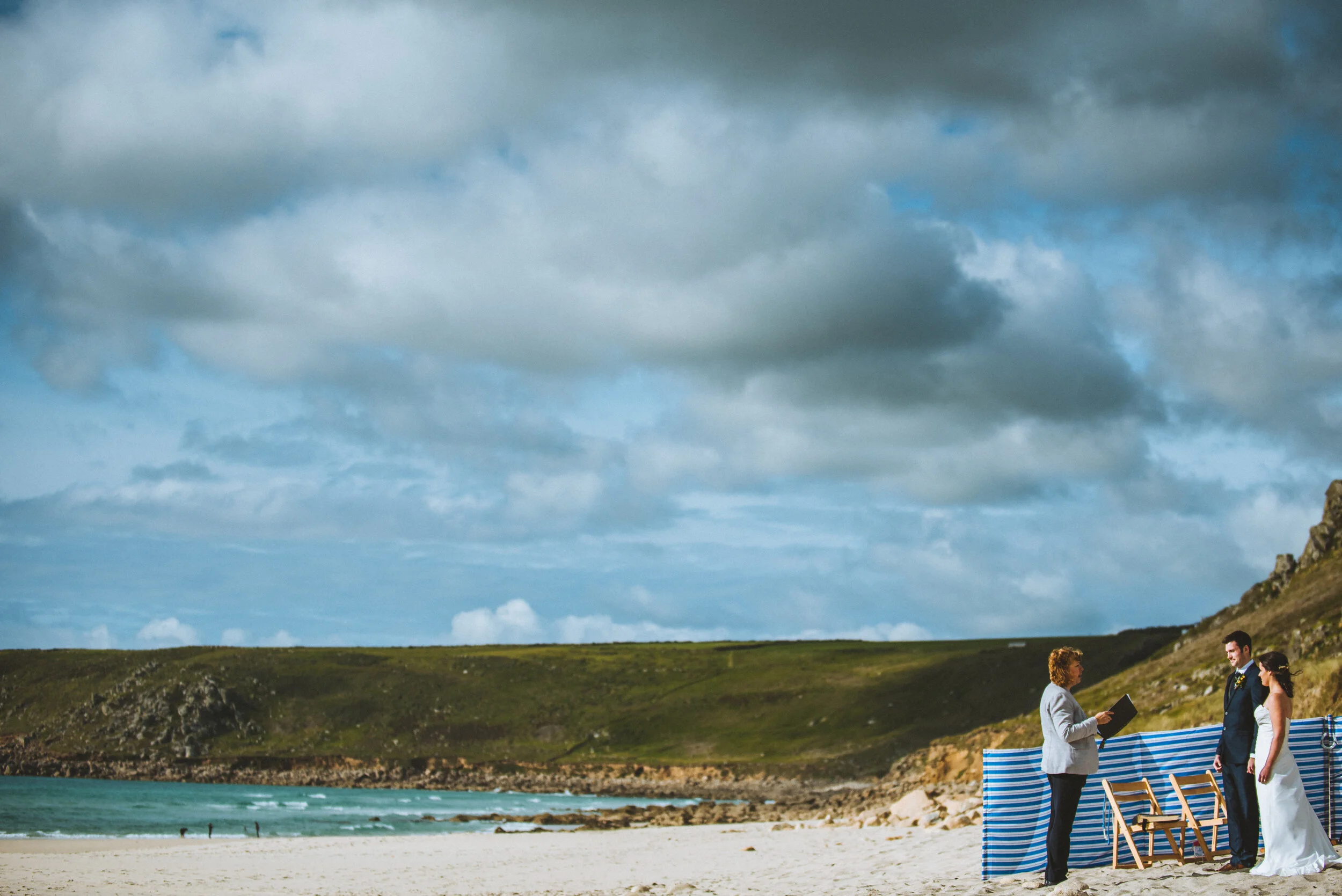 A wedding ceremony taking place on a beach with an officiant, bride, and groom in front of a blue and white striped backdrop, with rocky hills and a cloudy sky in the background.