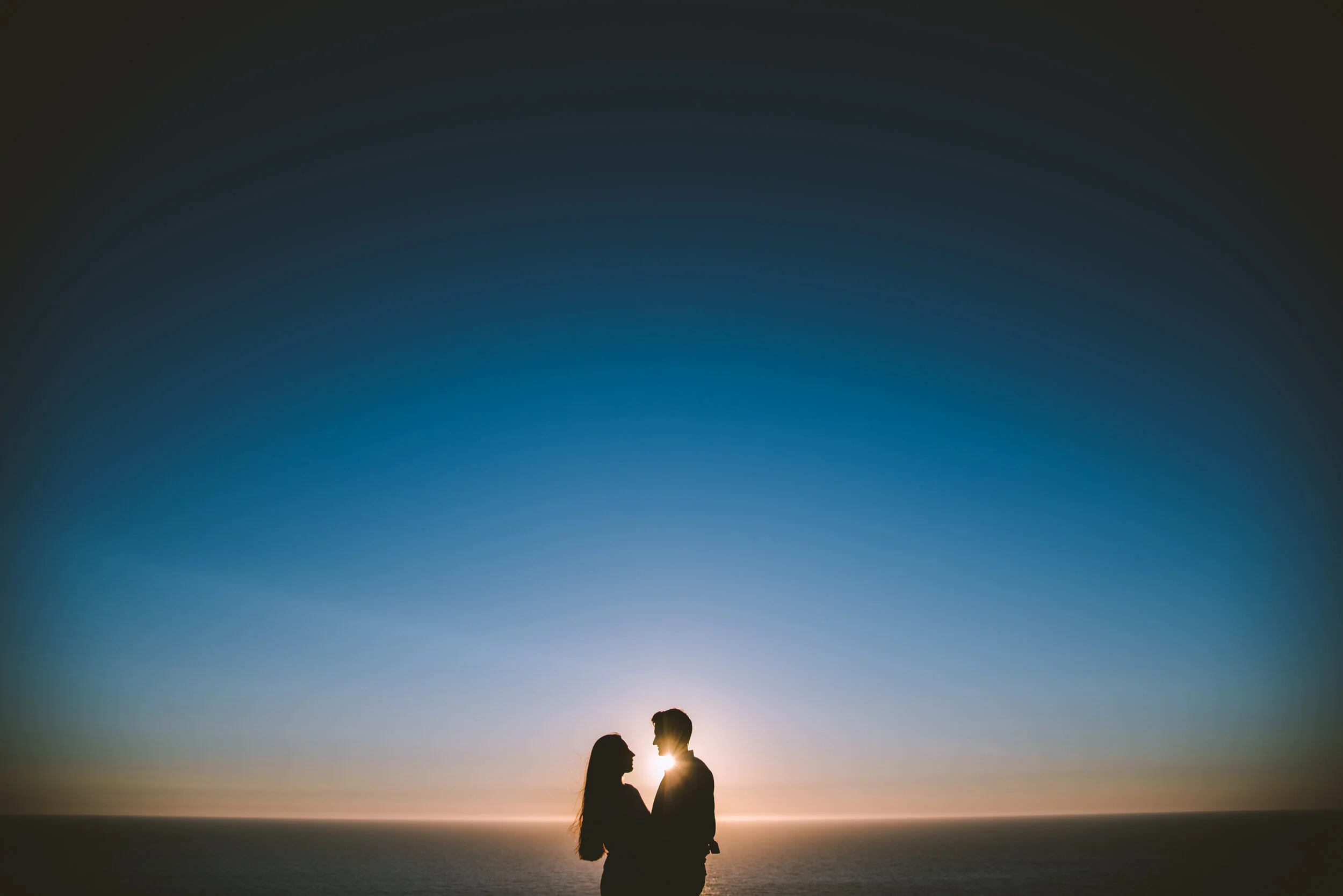 Silhouette of a couple standing close together during sunset by the ocean.