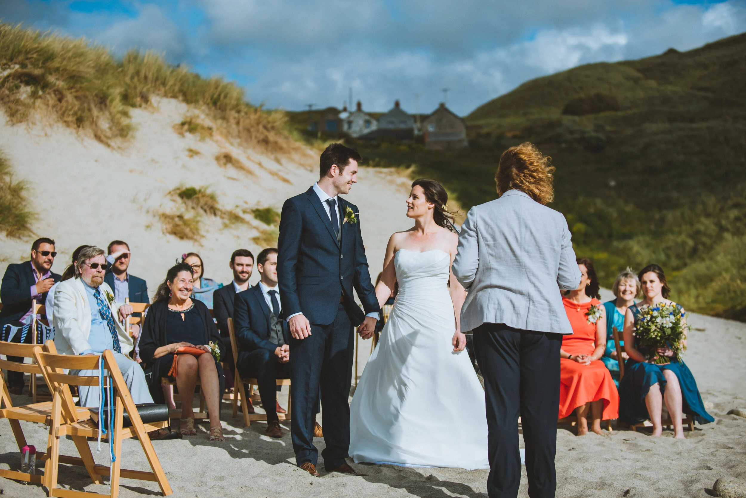 A wedding ceremony taking place on a sandy beach with grassy dunes in the background. The bride and groom are holding hands, facing a person officiating the ceremony. Guests are seated behind them, smiling and watching the ceremony.