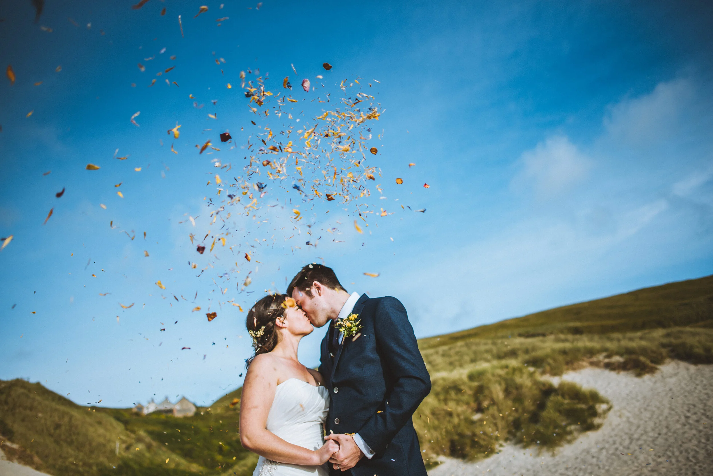 A bride and groom kiss outdoors under a blue sky with confetti in the air, standing on a grassy dune.