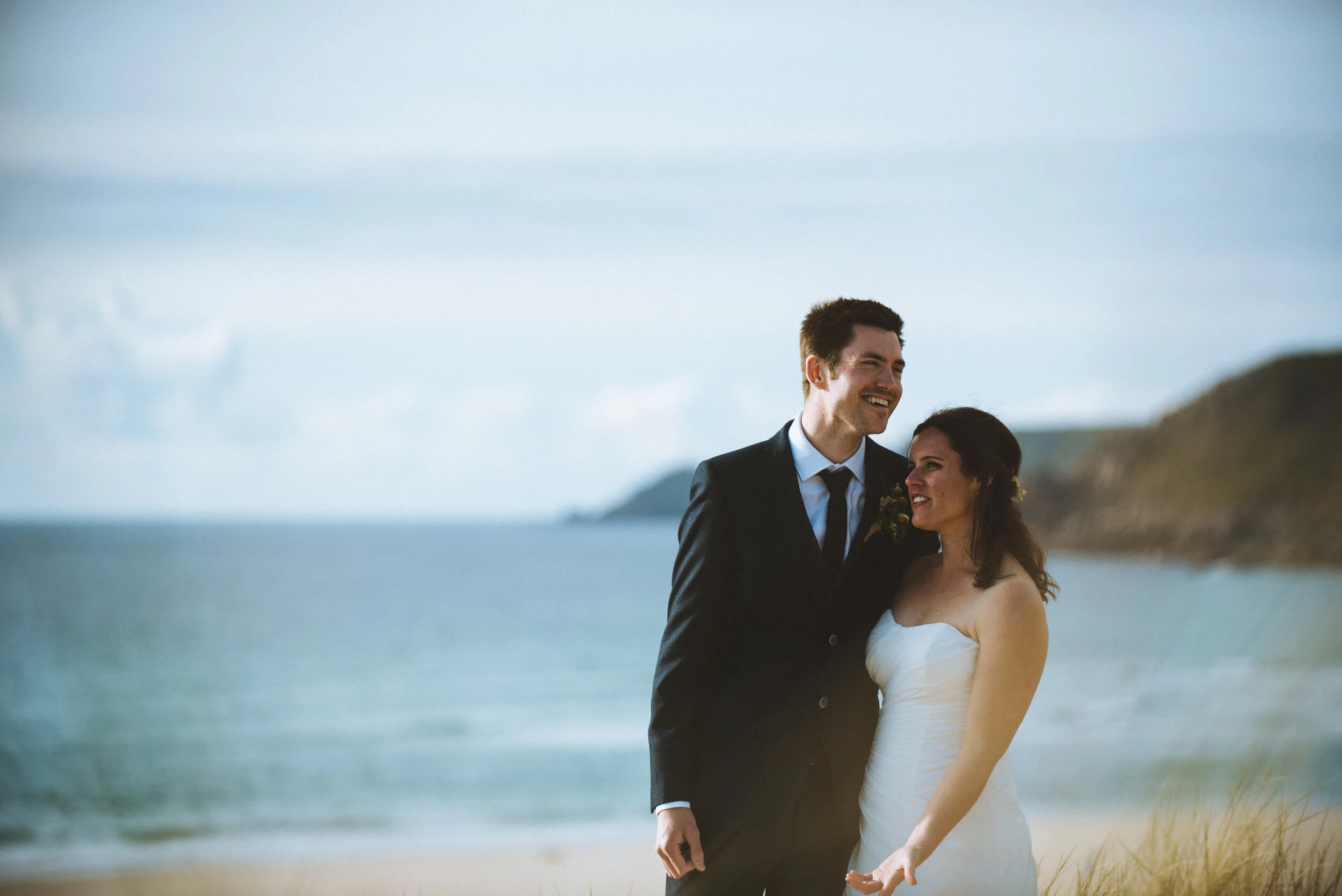 A bride and groom standing on a beach, smiling and enjoying each other's company, with ocean and cliffs in the background.