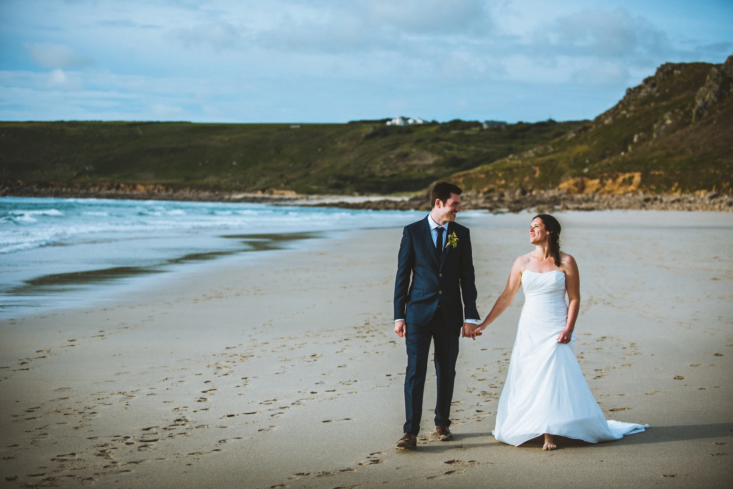 A bride and groom walking hand-in-hand on the beach, smiling at each other, with dunes and a cloudy sky in the background.