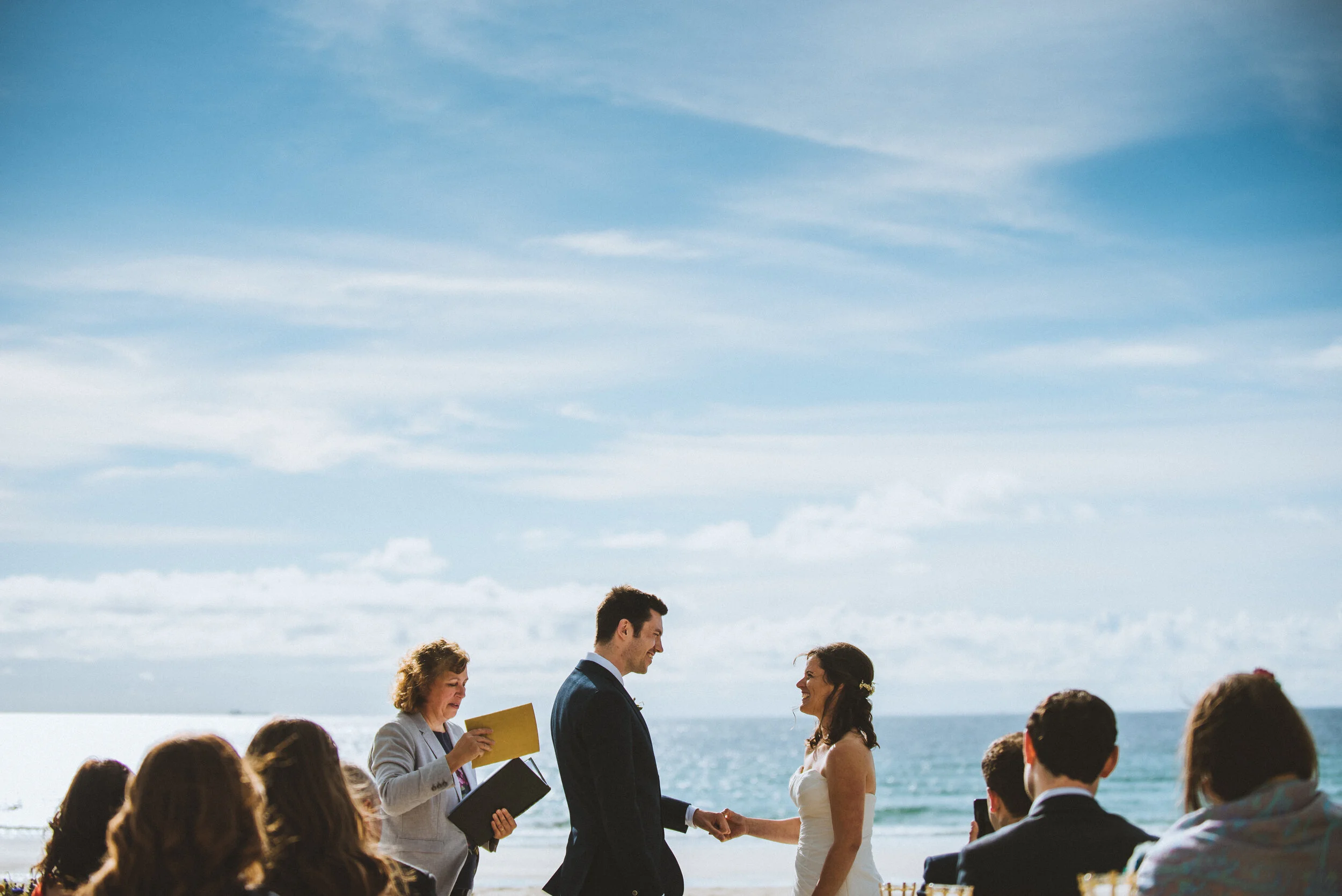A couple getting married on the beach, holding hands and smiling at each other, with an officiant reading from a book nearby and wedding guests sitting and standing on the sand, ocean and blue sky in the background.