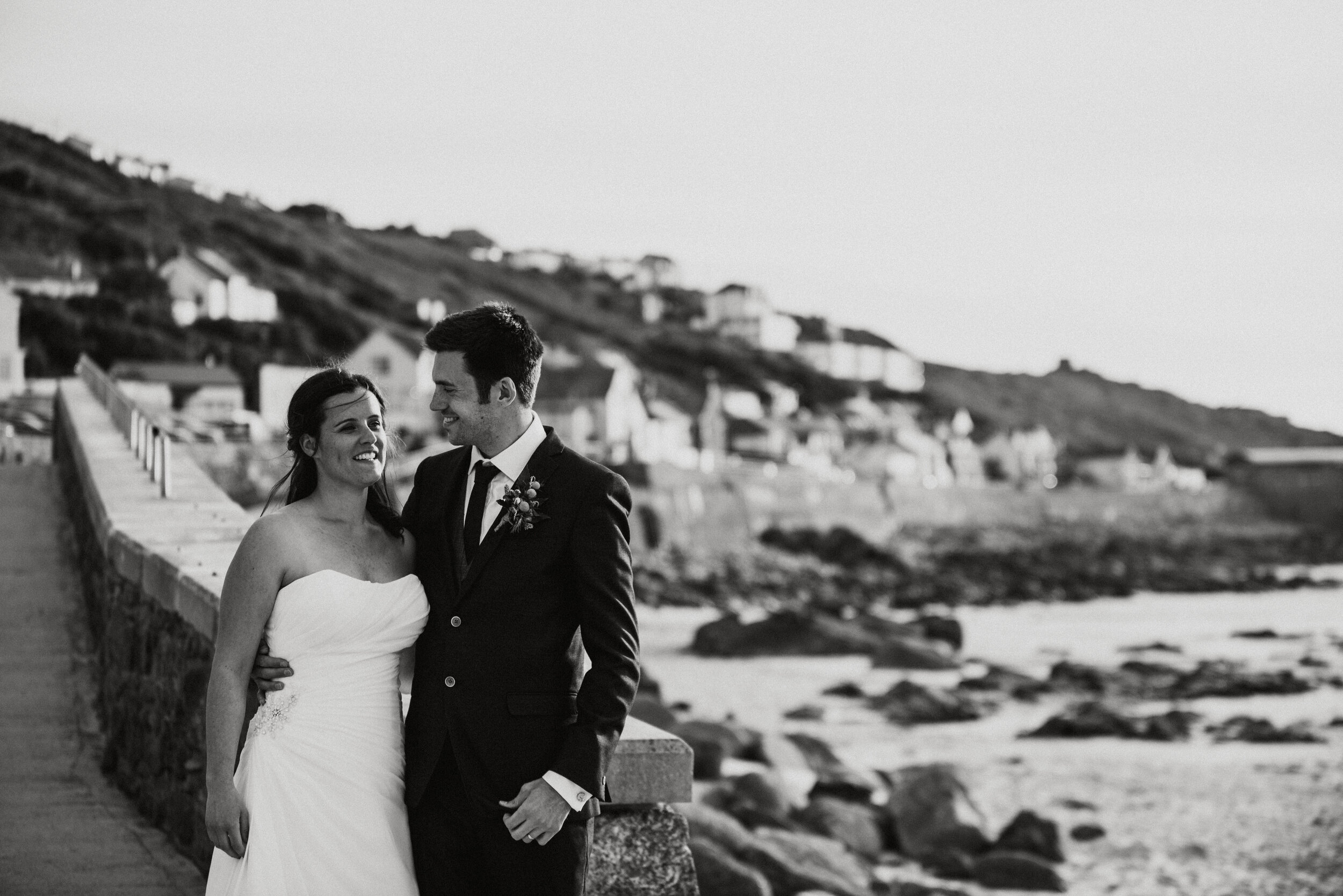 A bride and groom in wedding attire standing close together on a seaside promenade, smiling at each other with a rocky beach and houses on a hillside in the background.