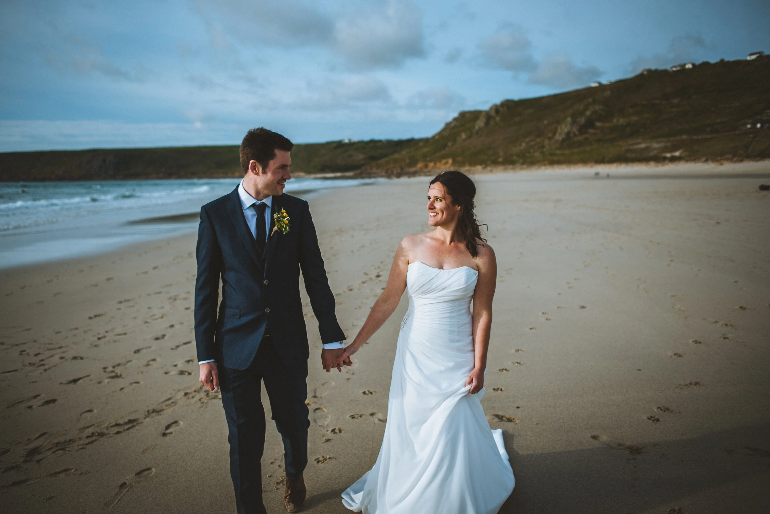 A couple in wedding attire walking hand in hand on a sandy beach with the ocean and cliffs in the background, under an overcast sky.