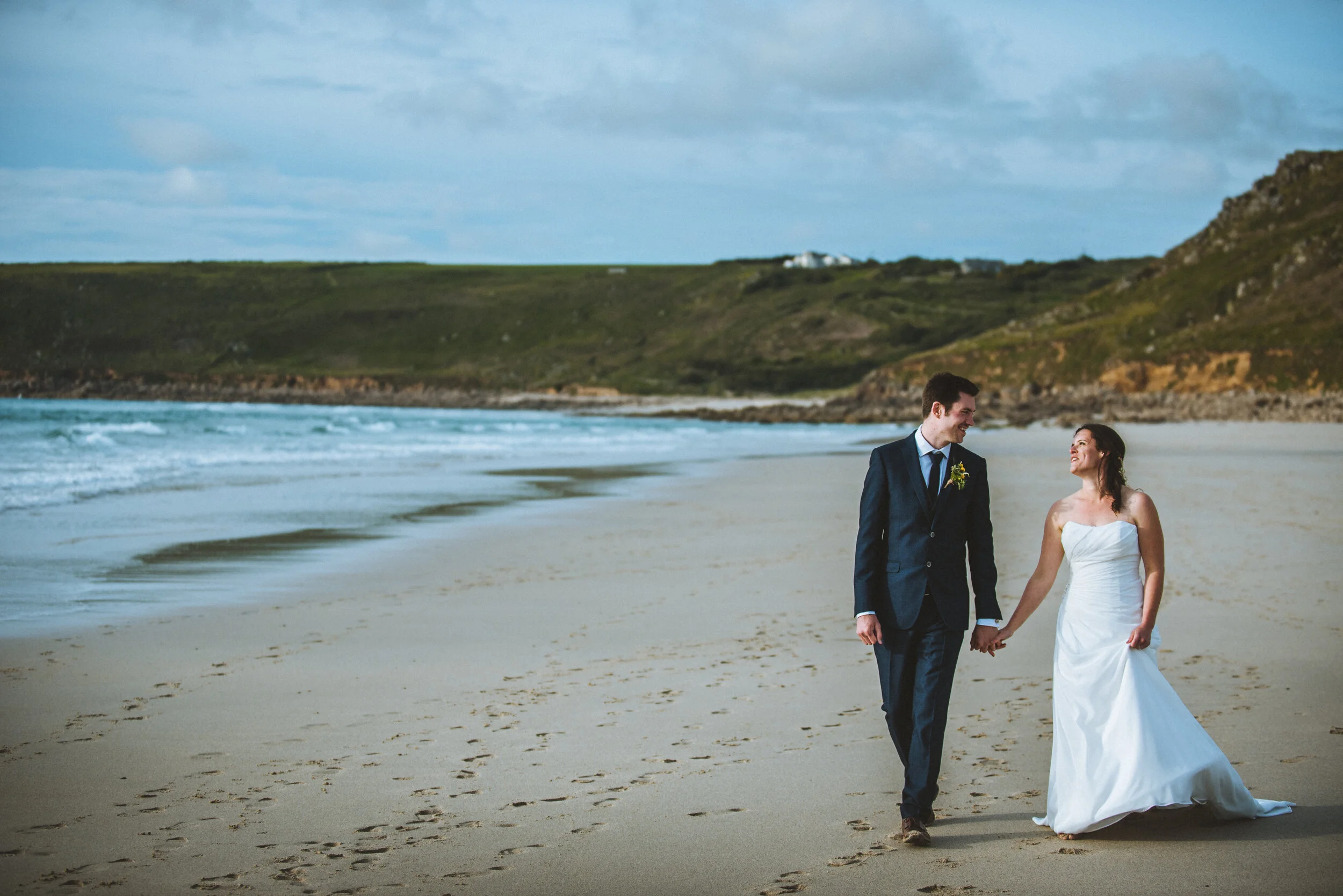 A couple in wedding attire walking hand in hand on a sandy beach with ocean waves and green hills in the background.