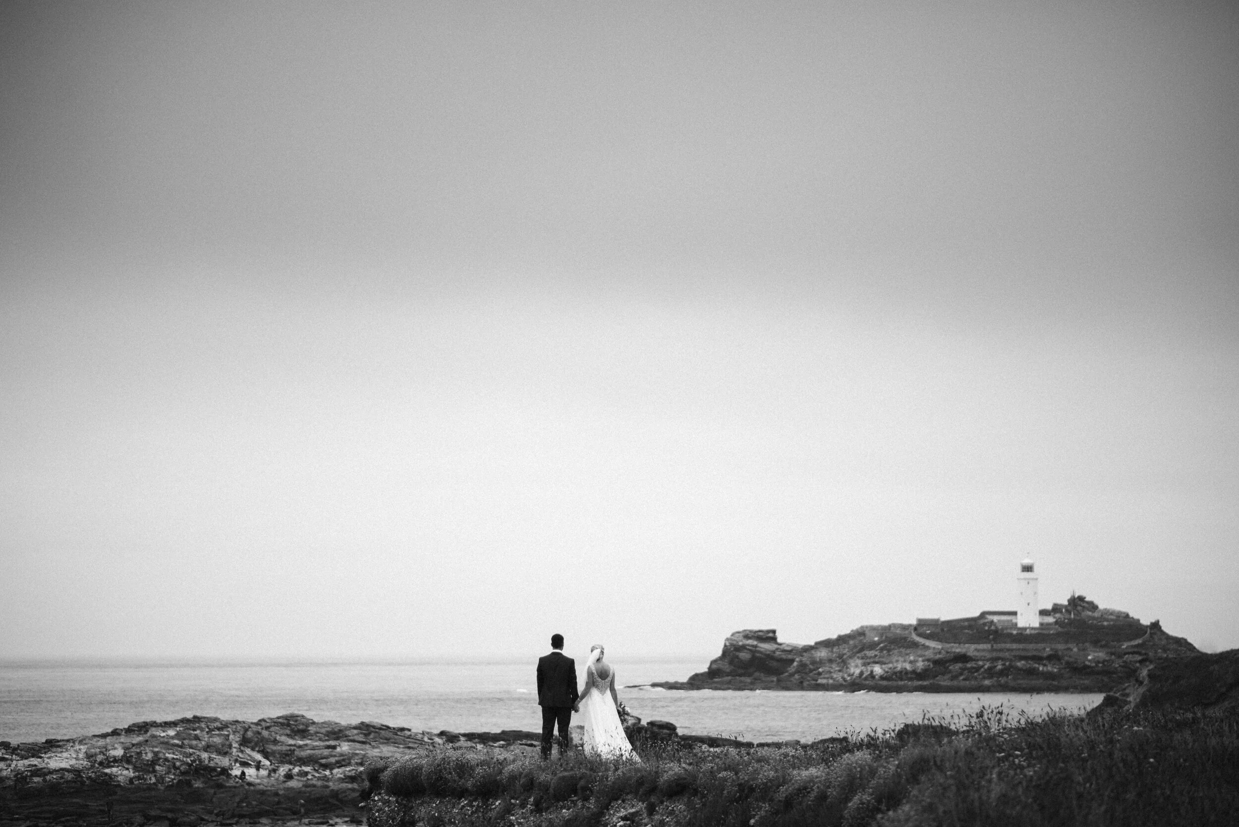 A black and white photo of a bride and groom holding hands near the shore, with a lighthouse in the distance on a rocky hill.