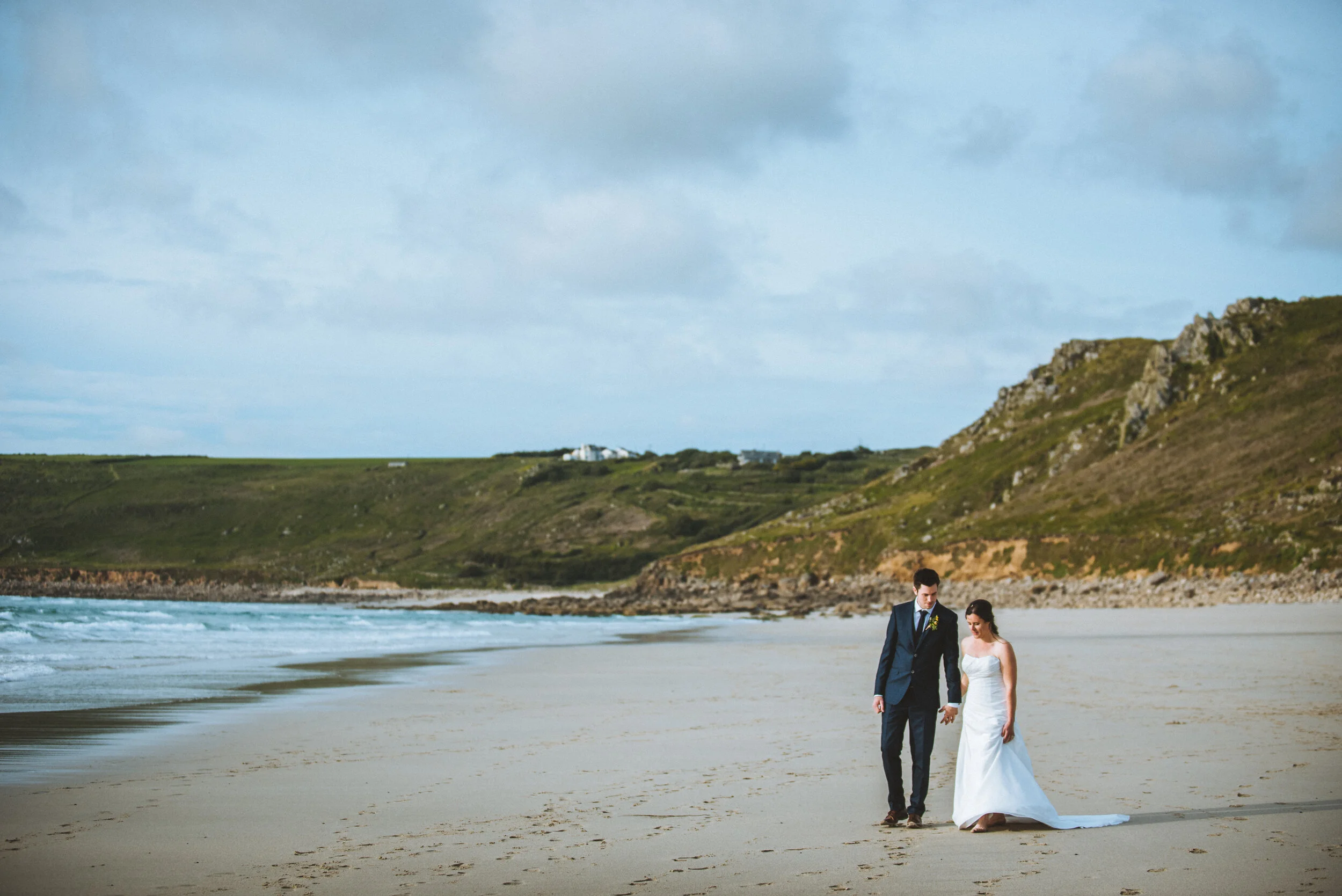 A bride and groom walking hand in hand on a beach with green hills in the background during daylight.