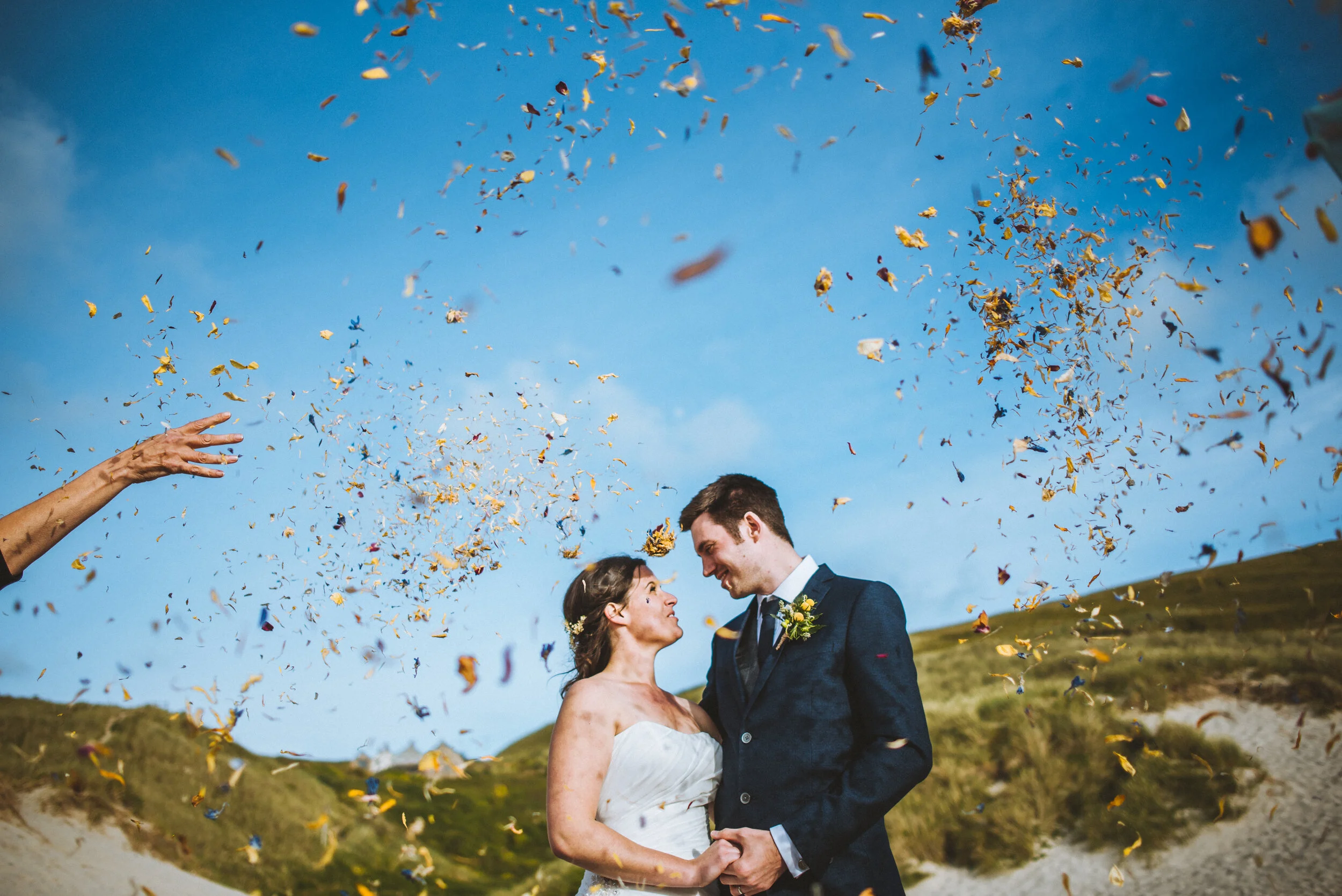A bride and groom standing close together outdoors, facing each other and holding hands, during their wedding celebration with confetti falling from above. The background shows grassy dunes and a blue sky.