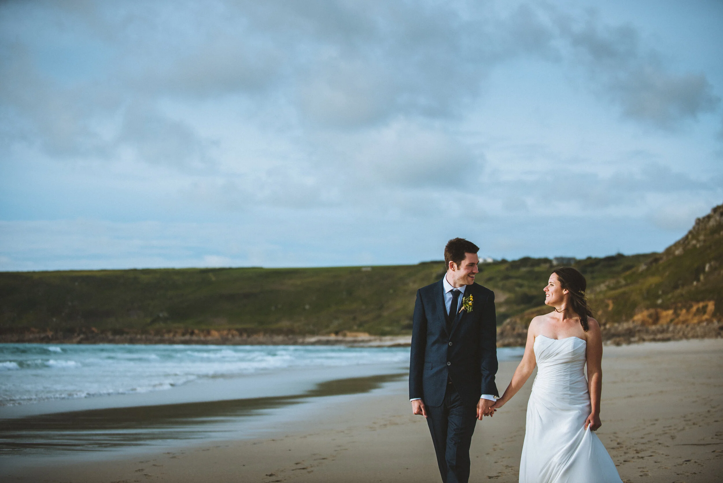 A happy couple, dressed in wedding attire, walking hand in hand on the beach during sunset or late afternoon, with ocean waves and cliffs in the background.