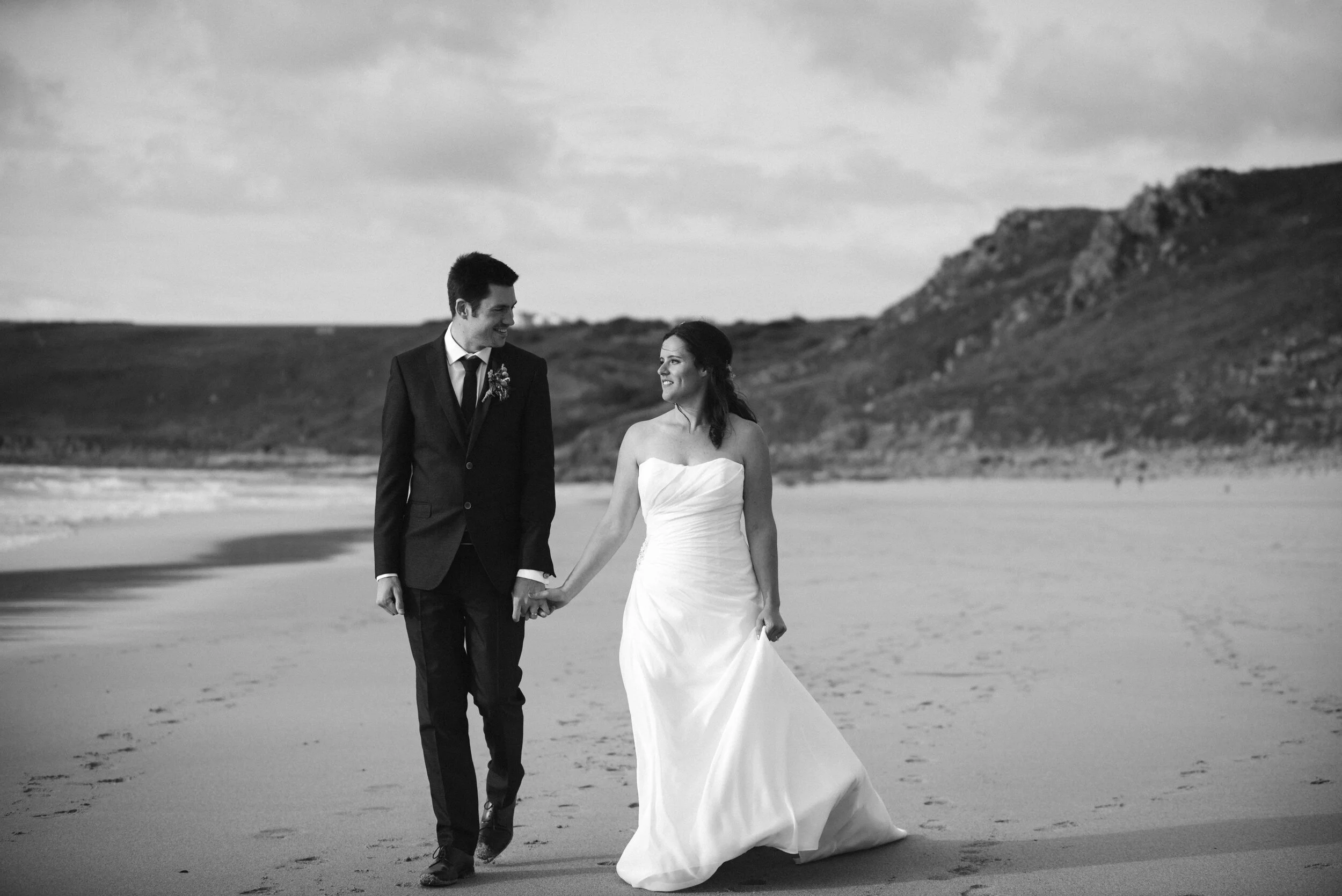 Black and white photo of a bride and groom walking hand in hand on a beach, smiling at each other, with cliffs and cloudy sky in the background.