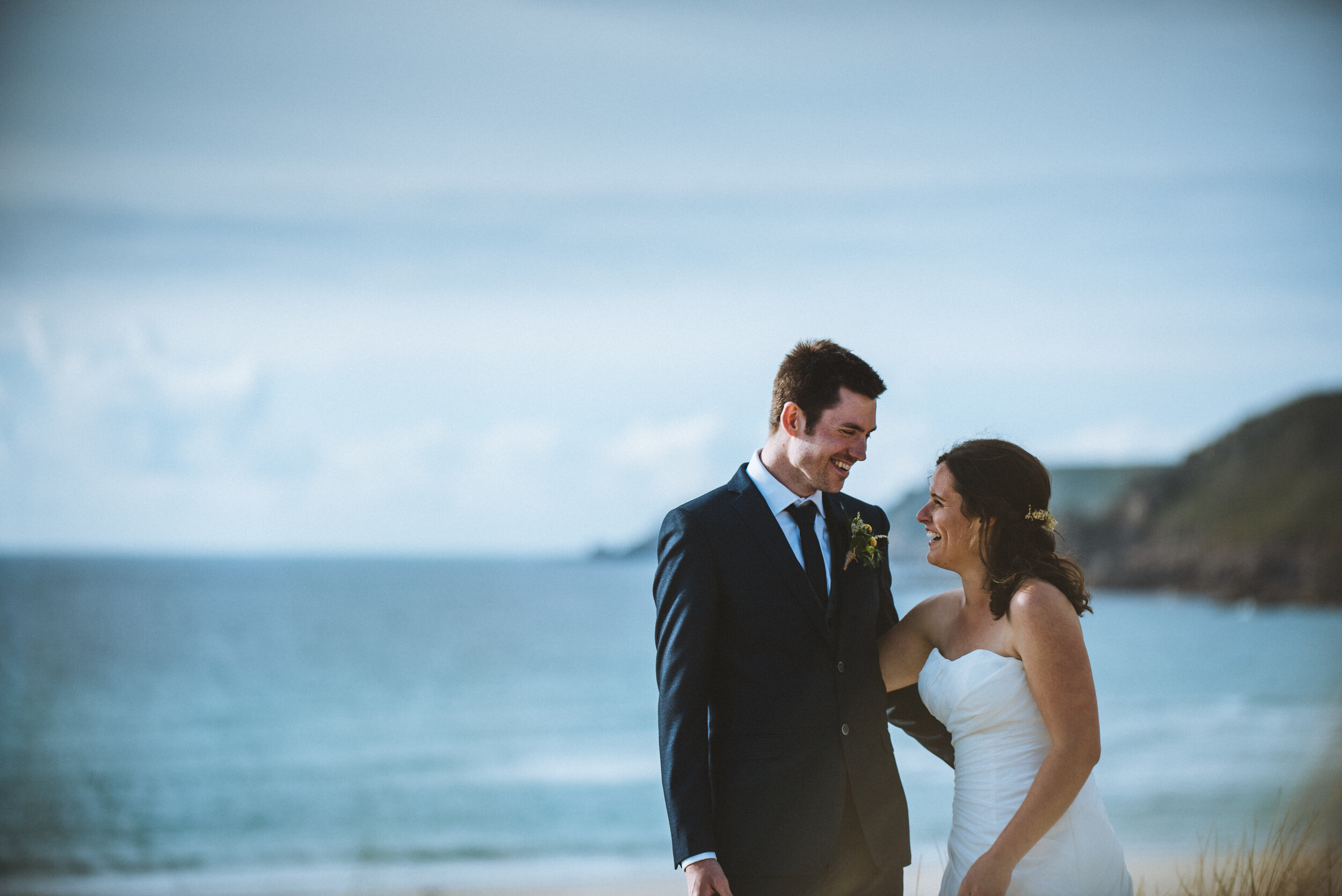 A newlywed couple standing on a beach, smiling at each other, with the ocean and sky in the background.