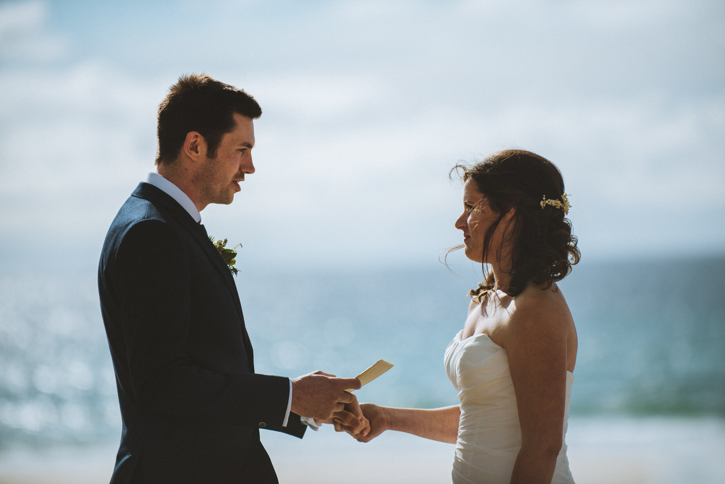 A man in a tuxedo and a woman in a wedding dress exchanging vows on the beach during a wedding ceremony.
