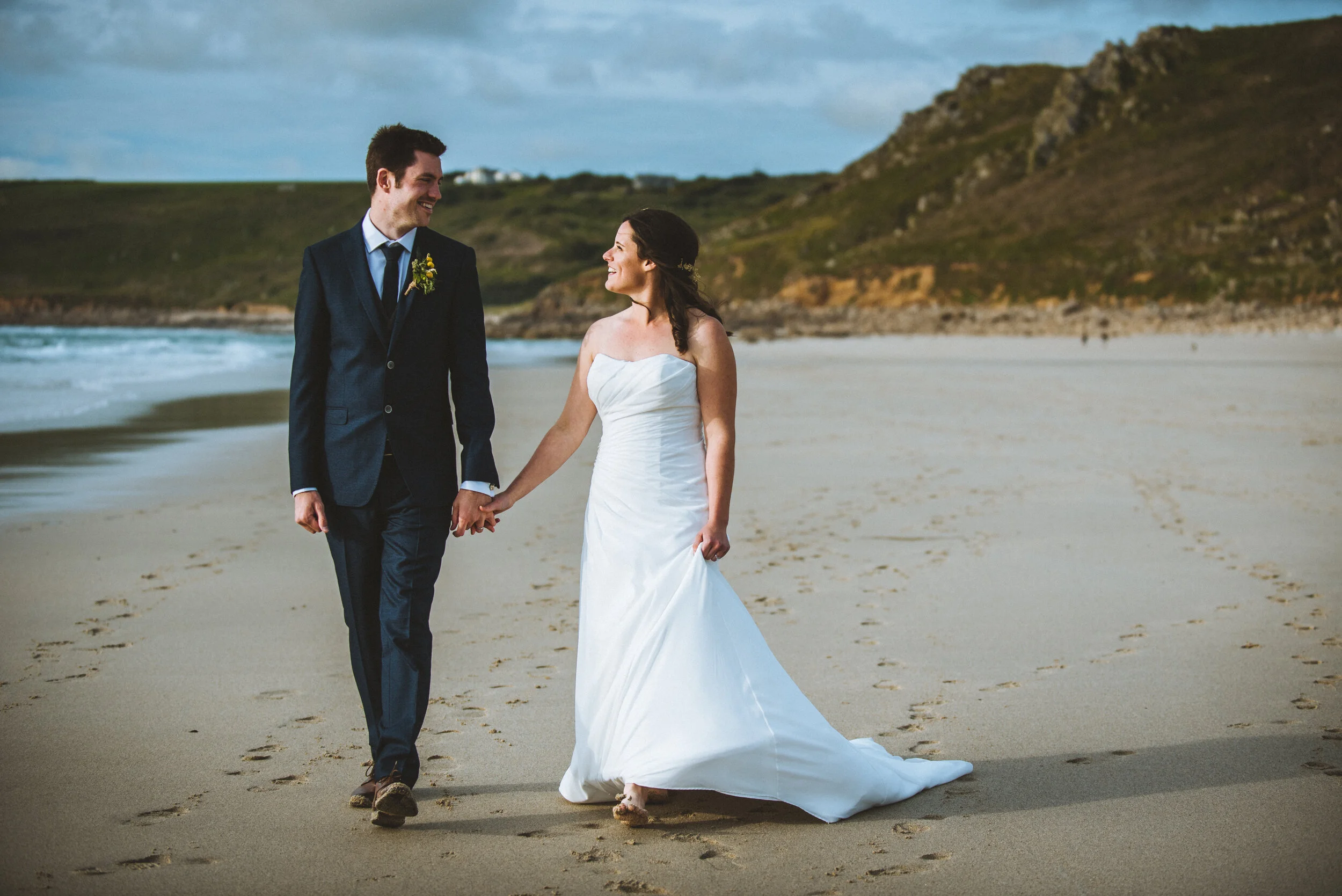 A bride and groom walking hand in hand on a beach, smiling at each other, with a rocky hillside in the background.