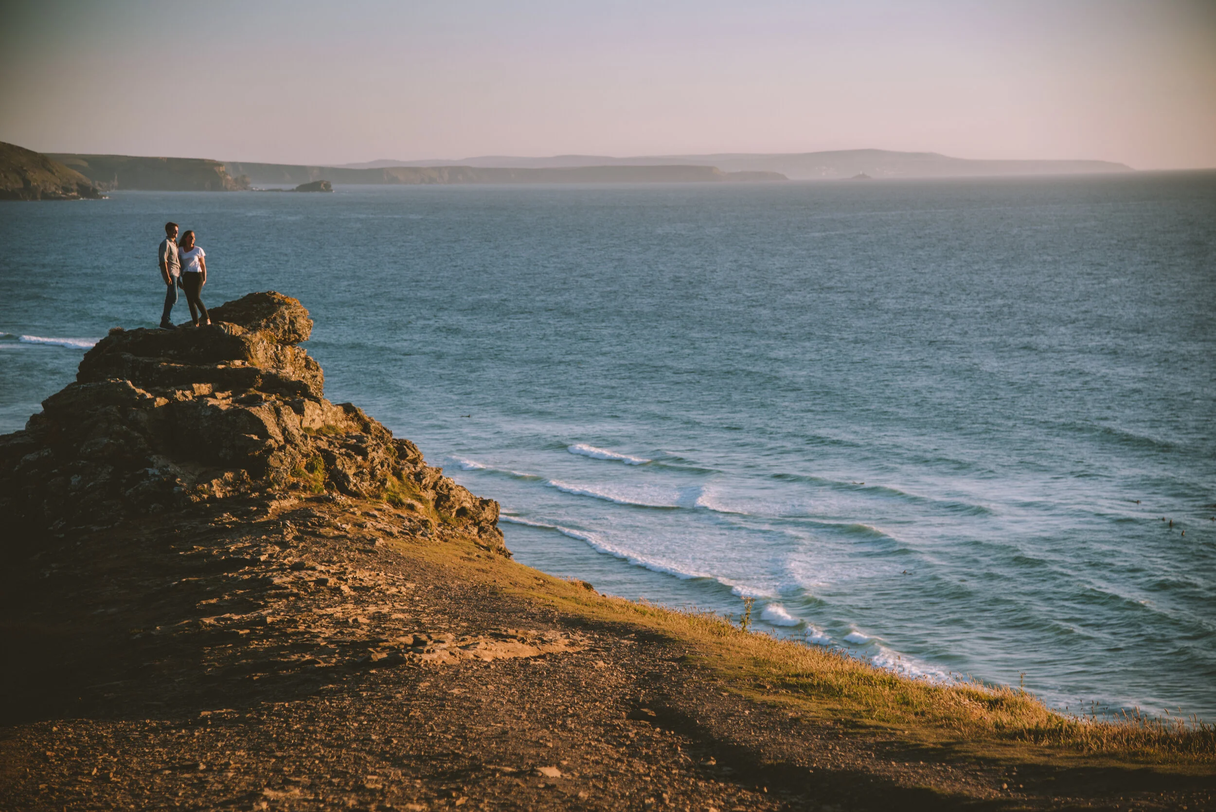 Two people standing on a rocky cliff overlooking the ocean during sunset.