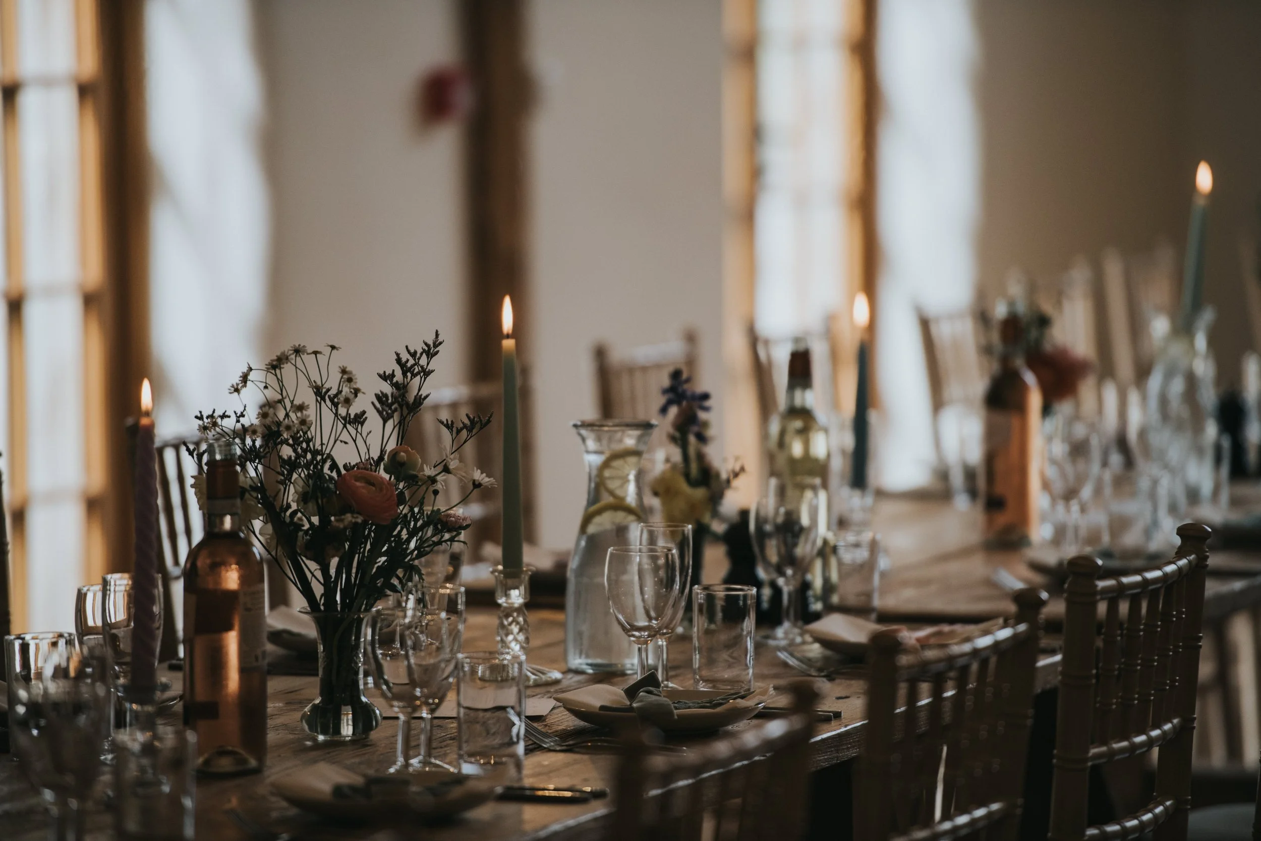 Elegant dining table set with flowers, candles, glasses, and cutlery near windows with soft natural lighting.