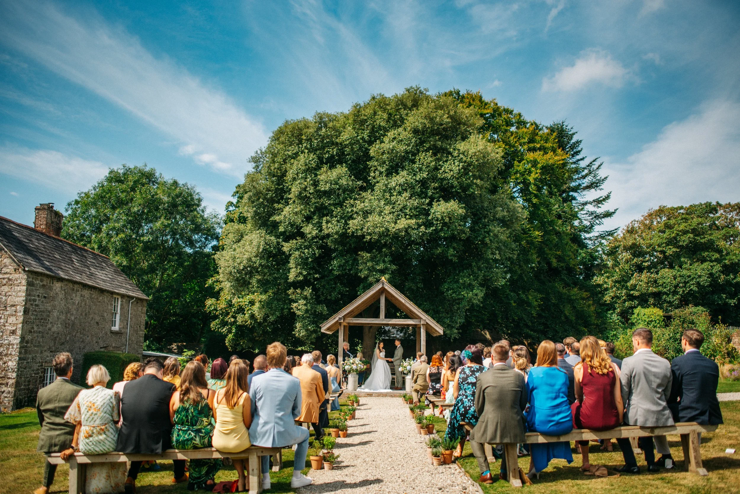 Outdoor wedding ceremony with guests seated on benches, couple standing under a wooden arch, large trees and partly cloudy sky in the background.