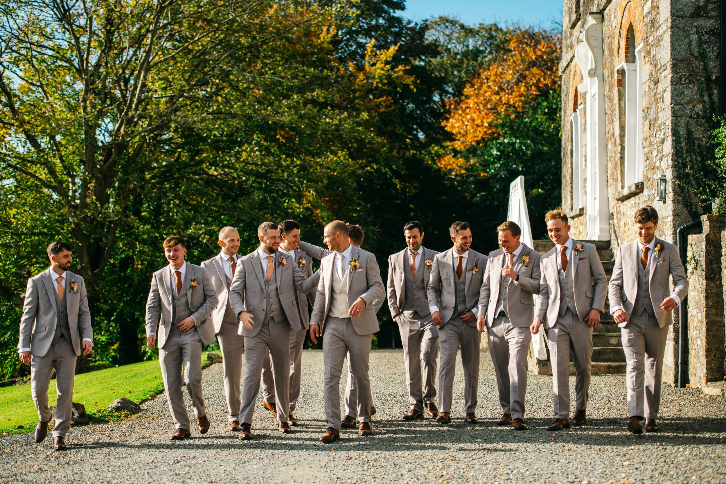 A group of men in gray suits with orange ties walking outdoors on a sunny day, wedding party, next to a stone building with large windows, trees with fall foliage in the background.