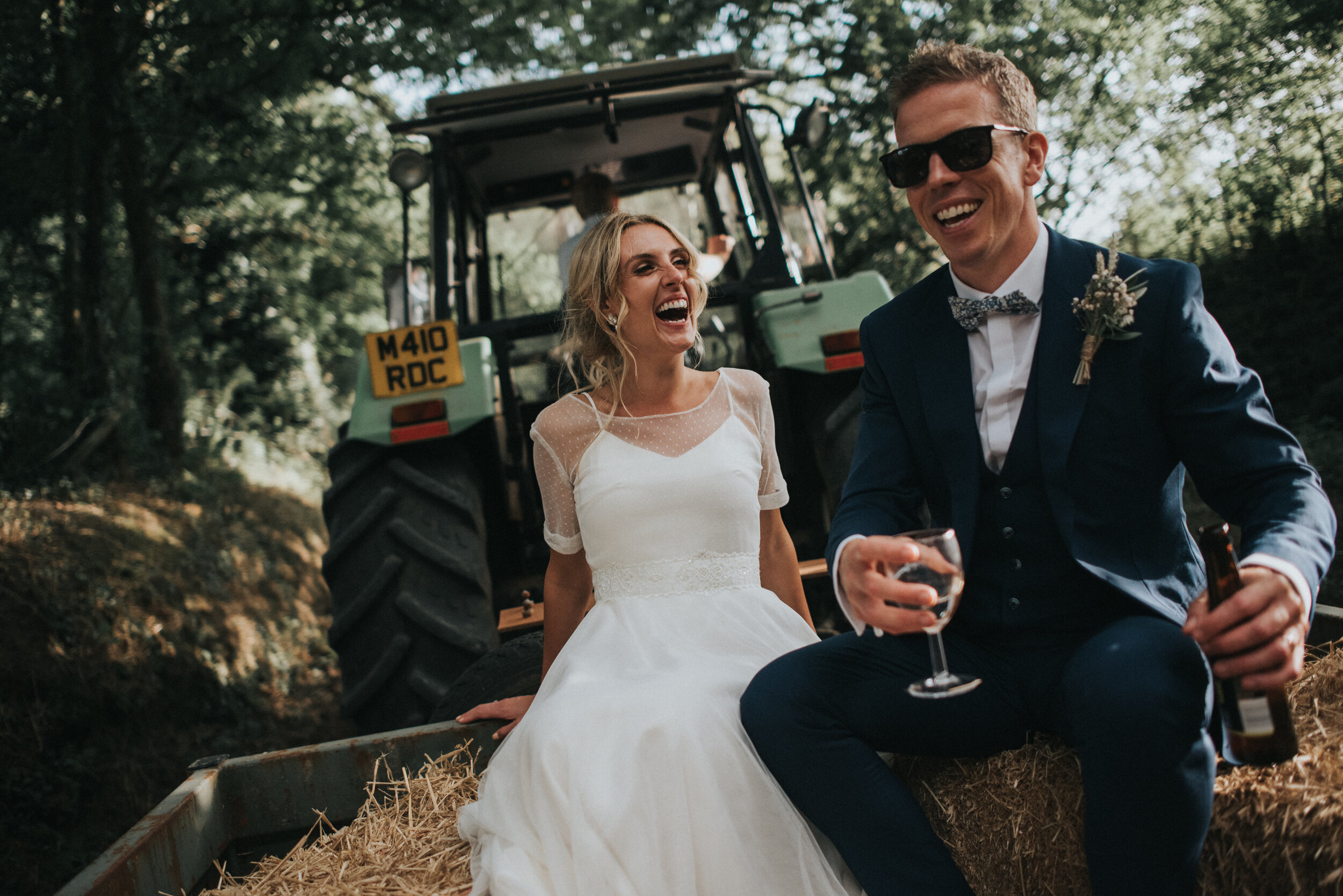 Bride and groom in wedding attire sitting on a hay bale in a wooded area, laughing and enjoying drinks, with a tractor in the background.