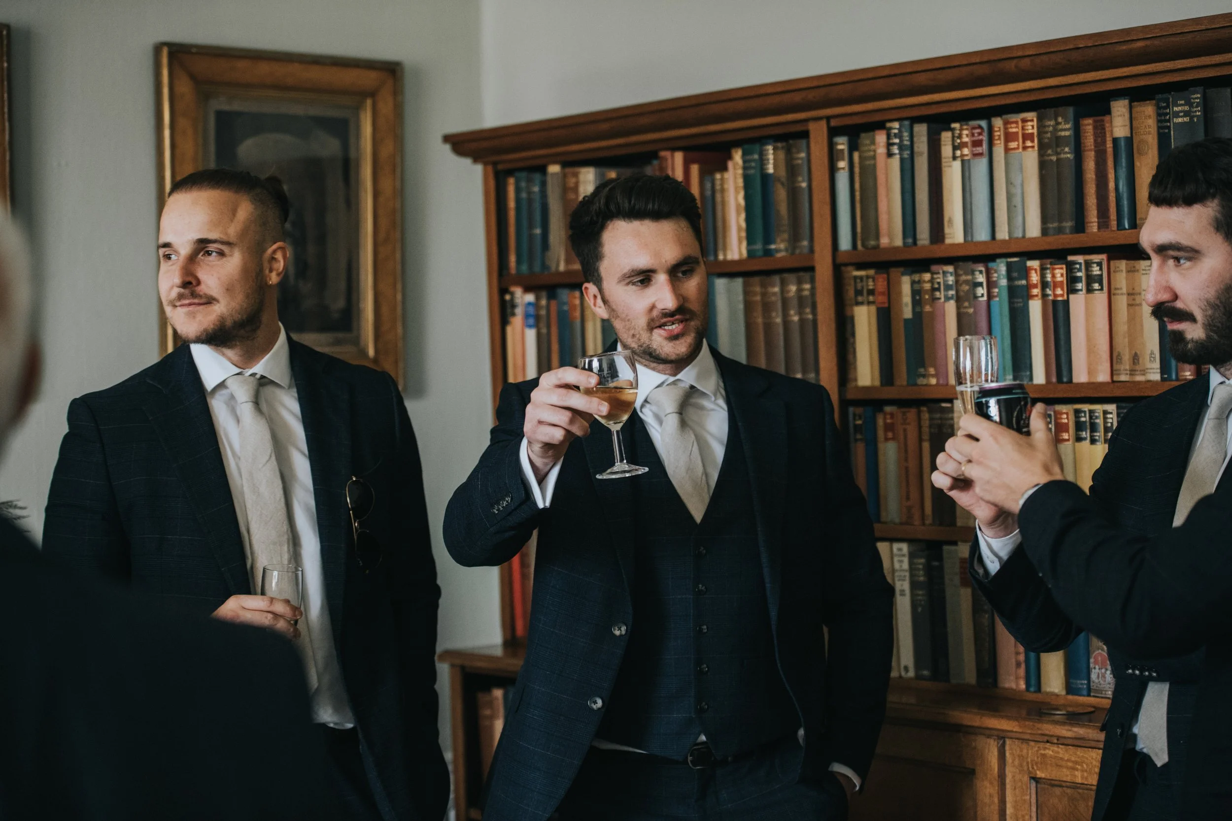 Three men dressed in suits having drinks and engaging in conversation in a library or study.
