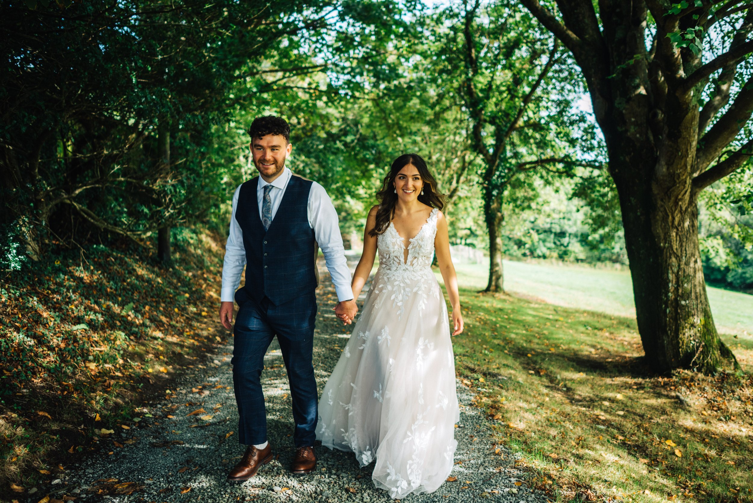 A bride and groom walking hand in hand on a tree-lined path during their wedding day.
