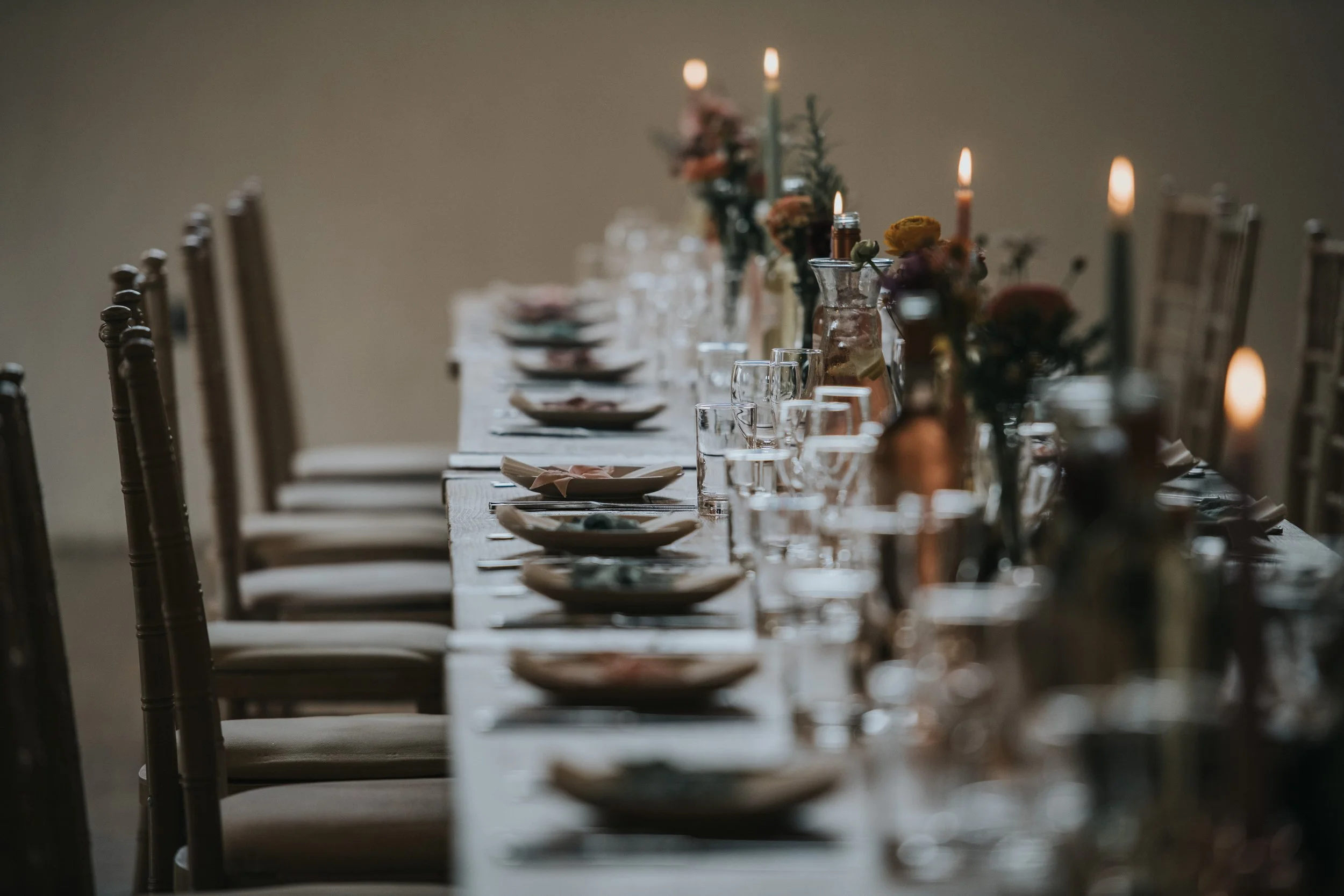 A long dinner table set with plates, glasses, napkins, and floral centerpieces with candles, in a softly lit room.