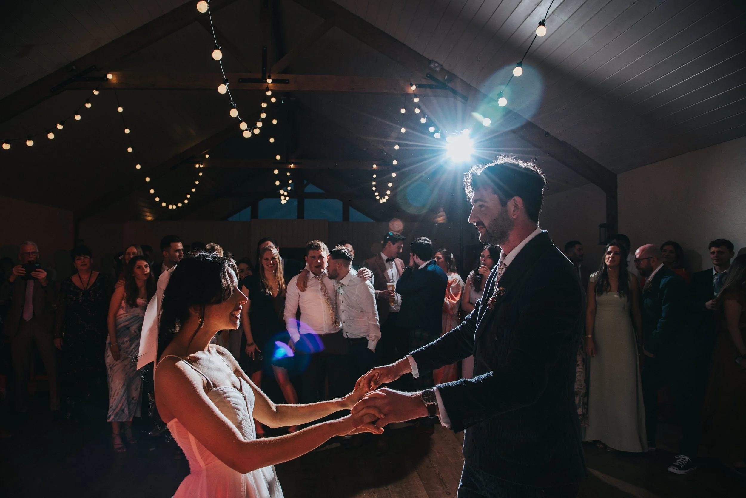 A bride and groom dancing at a wedding reception with guests watching, string lights hanging from the ceiling, and a bright light behind them creating a silhouette.