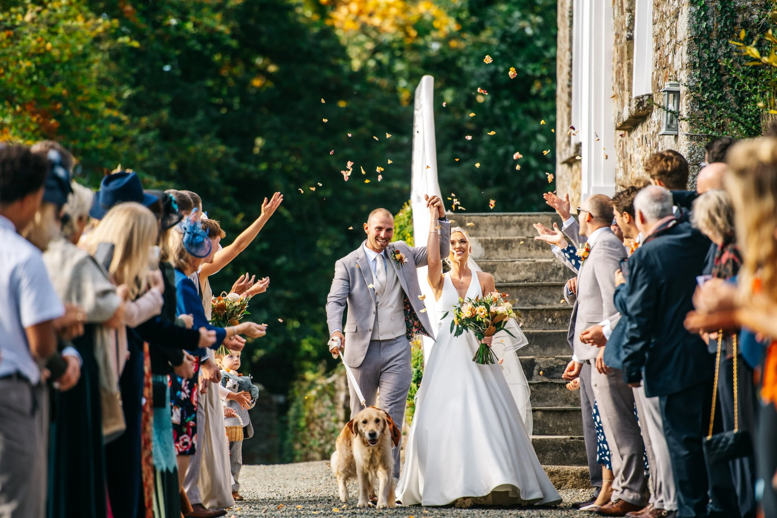 A newlywed couple celebrating their wedding outside, with the groom and bride holding hands and smiling while surrounded by guests throwing confetti. The groom is dressed in a light gray suit, and the bride is wearing a white wedding gown and holding