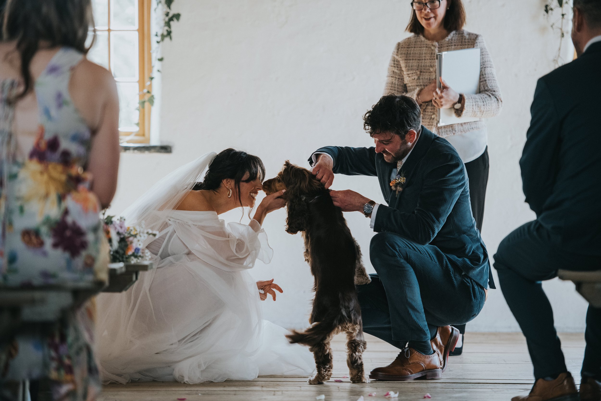 Bride and groom kneeling, smiling, and petting a dog during a wedding ceremony, with an officiant standing in the background and guests around them.