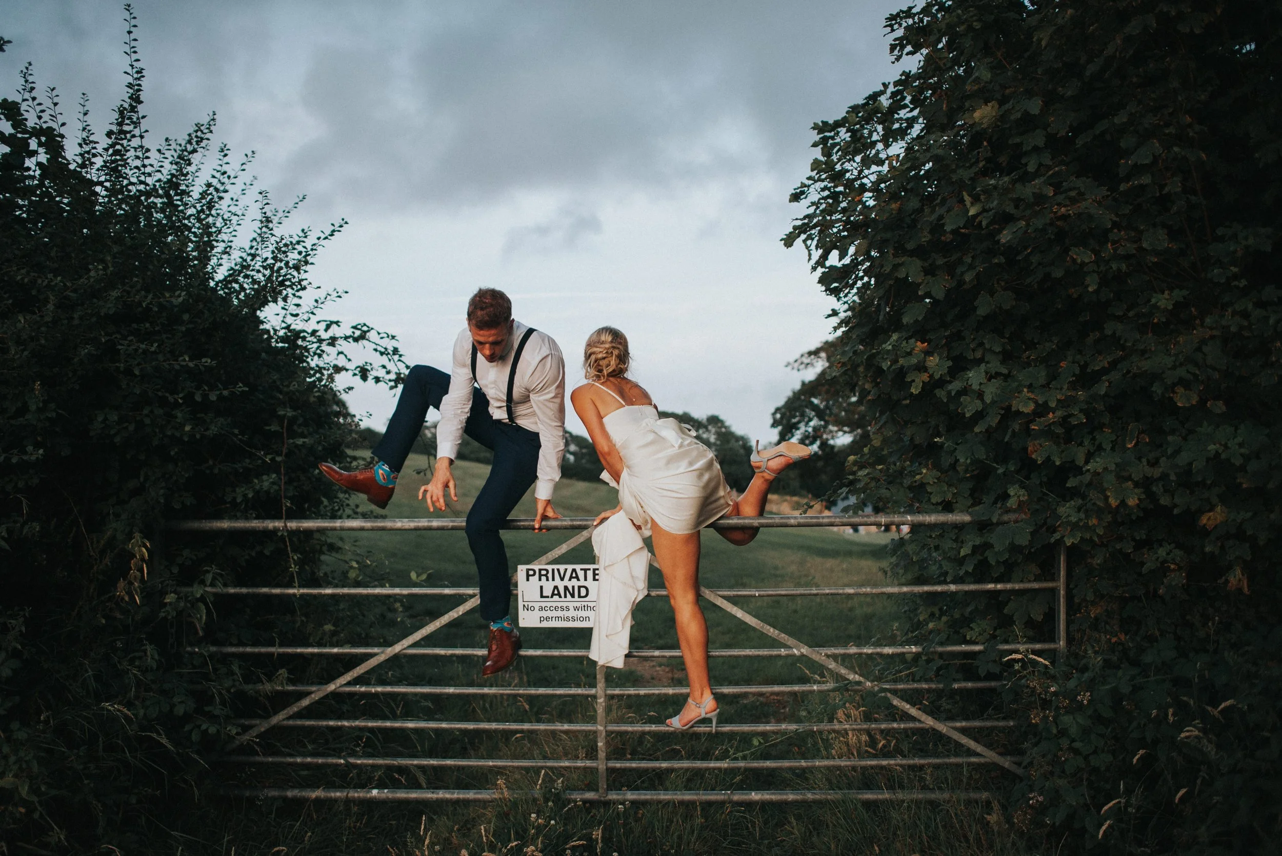 A man and woman dressed in formal attire sitting on a metal gate, with the man hanging off the gate and the woman resting her leg on the gate. The scene is outdoors with trees and a cloudy sky in the background.