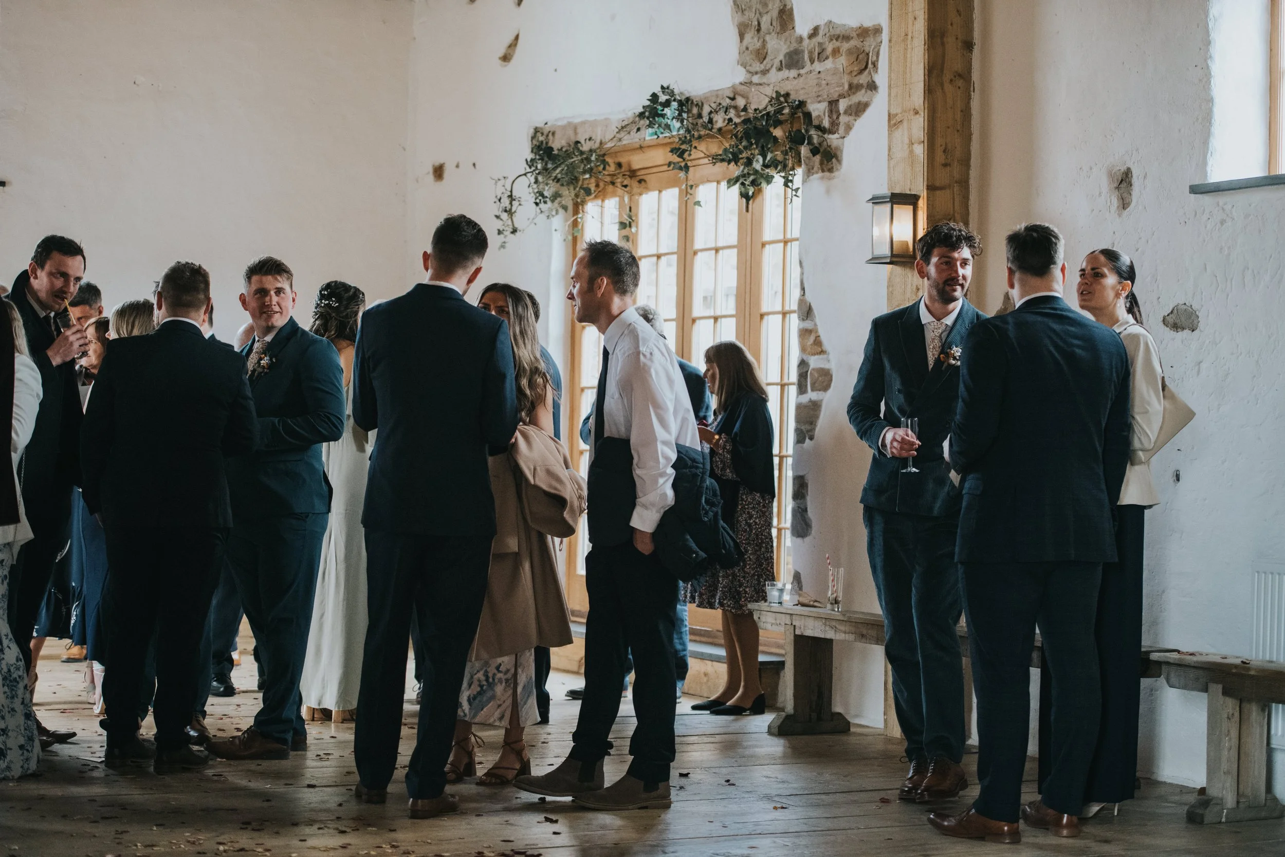 Group of people dressed in formal attire socializing at an indoor event, likely a wedding reception, with wooden floors, large windows, and a rustic decor.