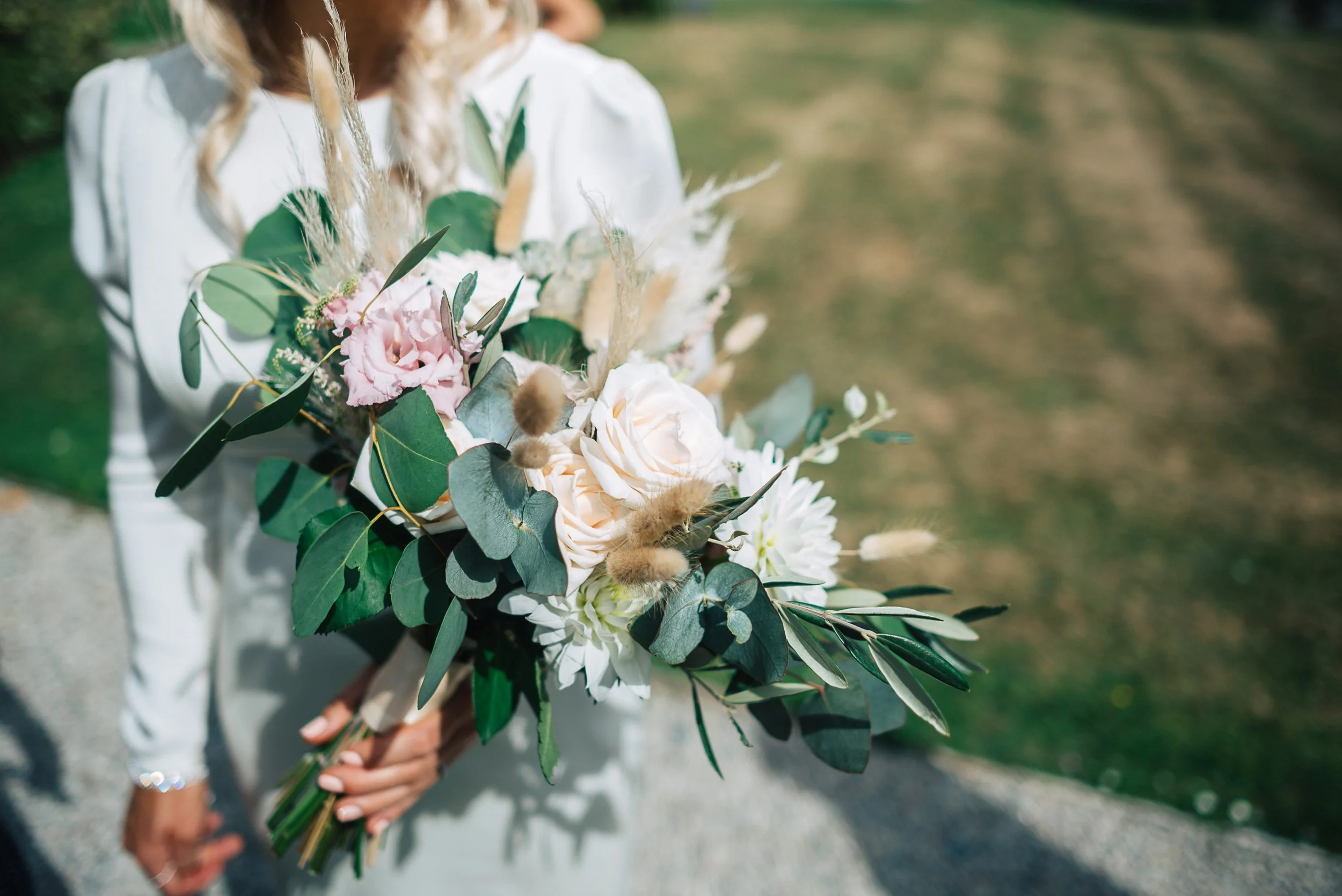 A person holding a bouquet of mixed flowers including white roses, pink flowers, greenery, and dried elements, outdoors on a sunny day.
