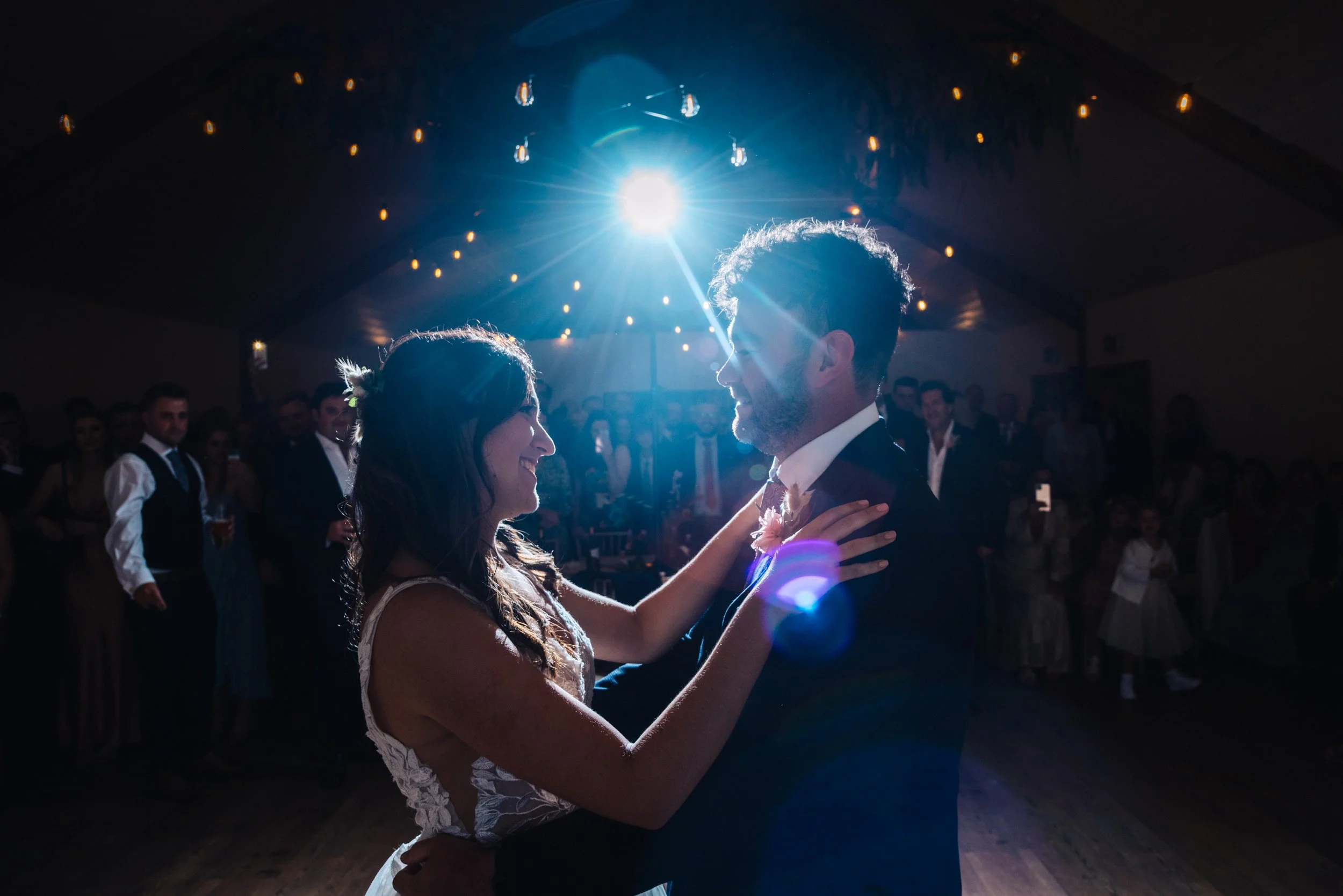 A bride and groom sharing their first dance at a wedding reception in a dimly lit hall with string lights, with guests watching in the background and a bright light shining down on them.