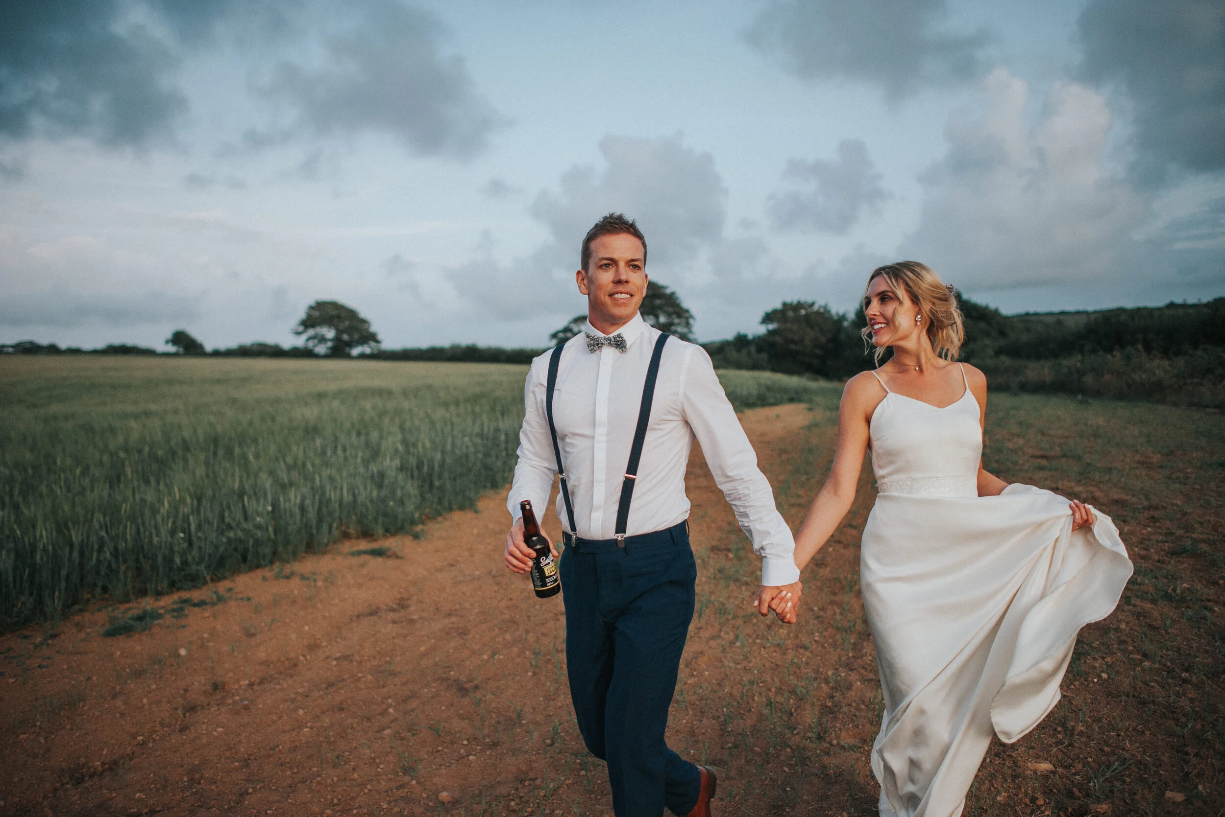 A man and woman in wedding attire holding hands and walking on a dirt path through a green field with trees and cloudy sky in the background.