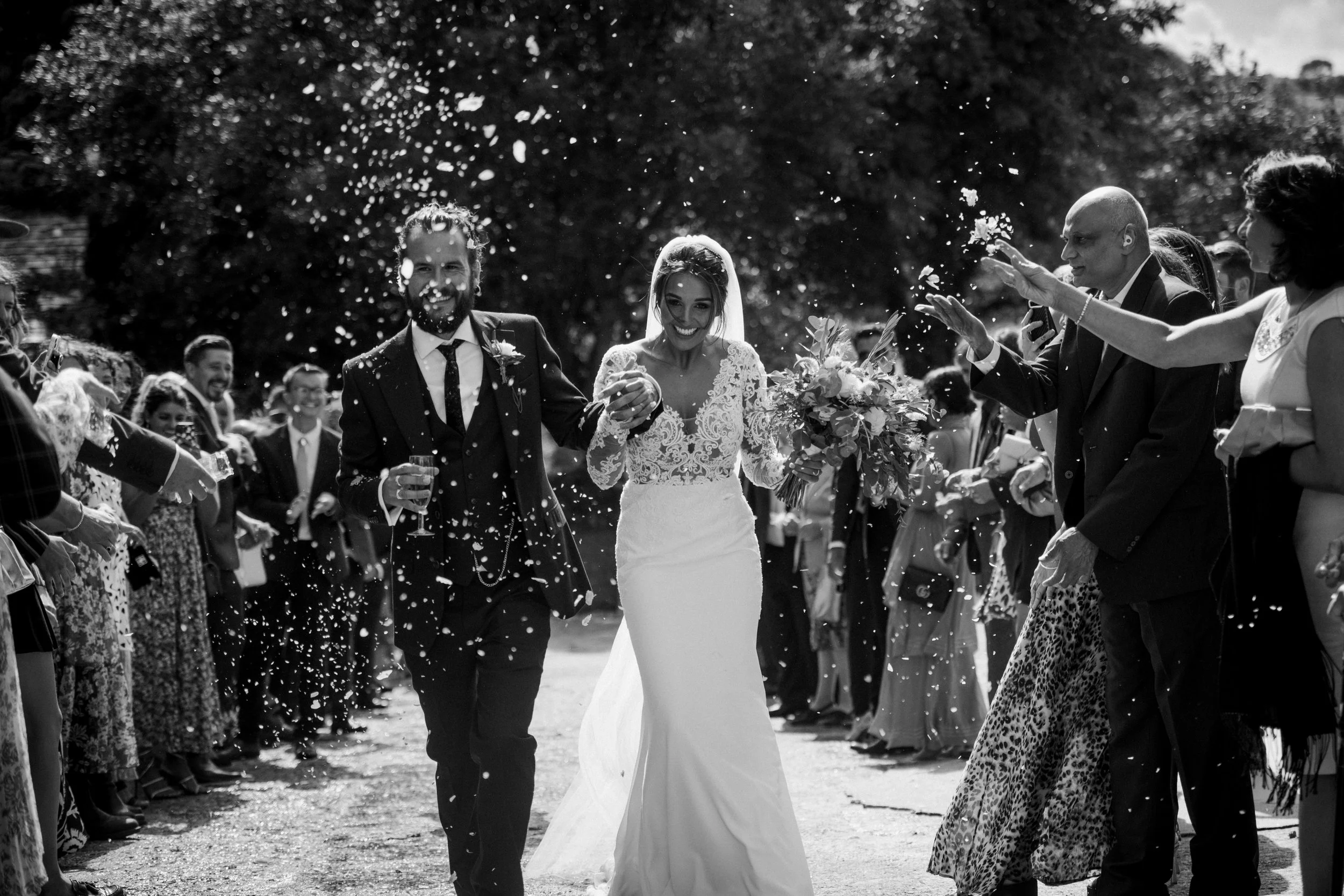 A bride and groom walk through a crowd of wedding guests throwing confetti during daytime outdoor wedding celebration.