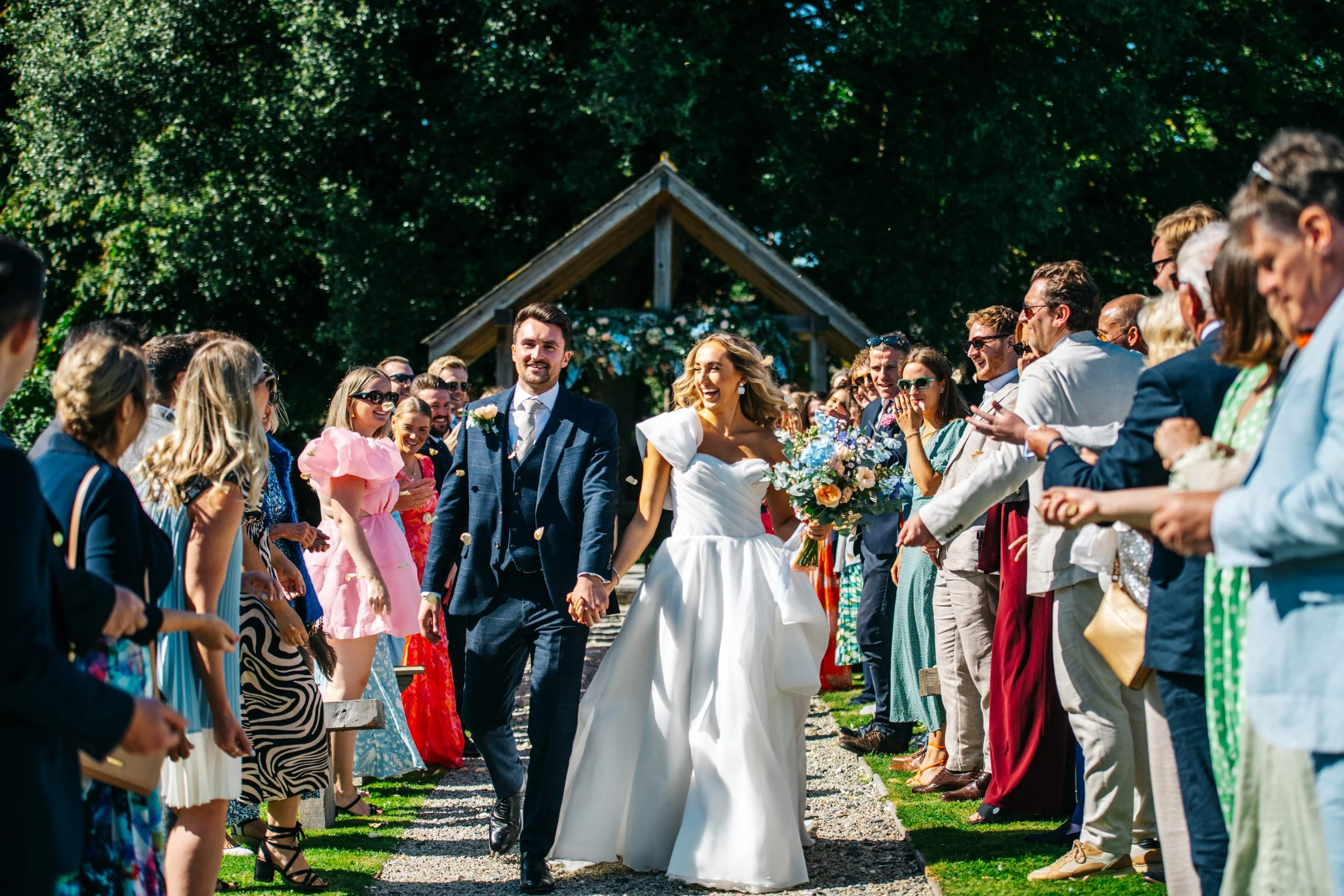 A newlywed couple walking through a crowd of celebrating guests outdoors on a sunny day, with greenery and a wooden structure in the background.