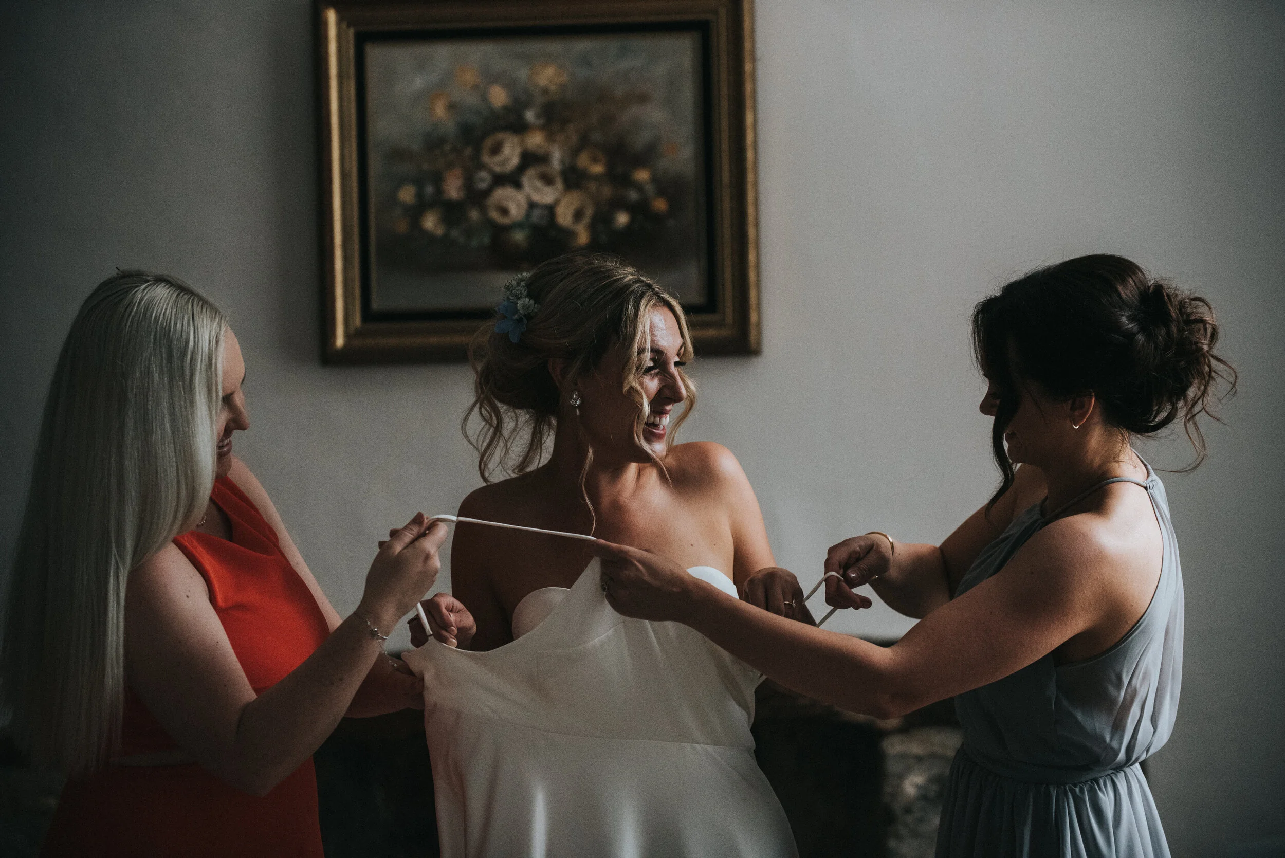 Three women, one in a wedding dress, preparing for a wedding, smiling and helping each other in a softly lit room.