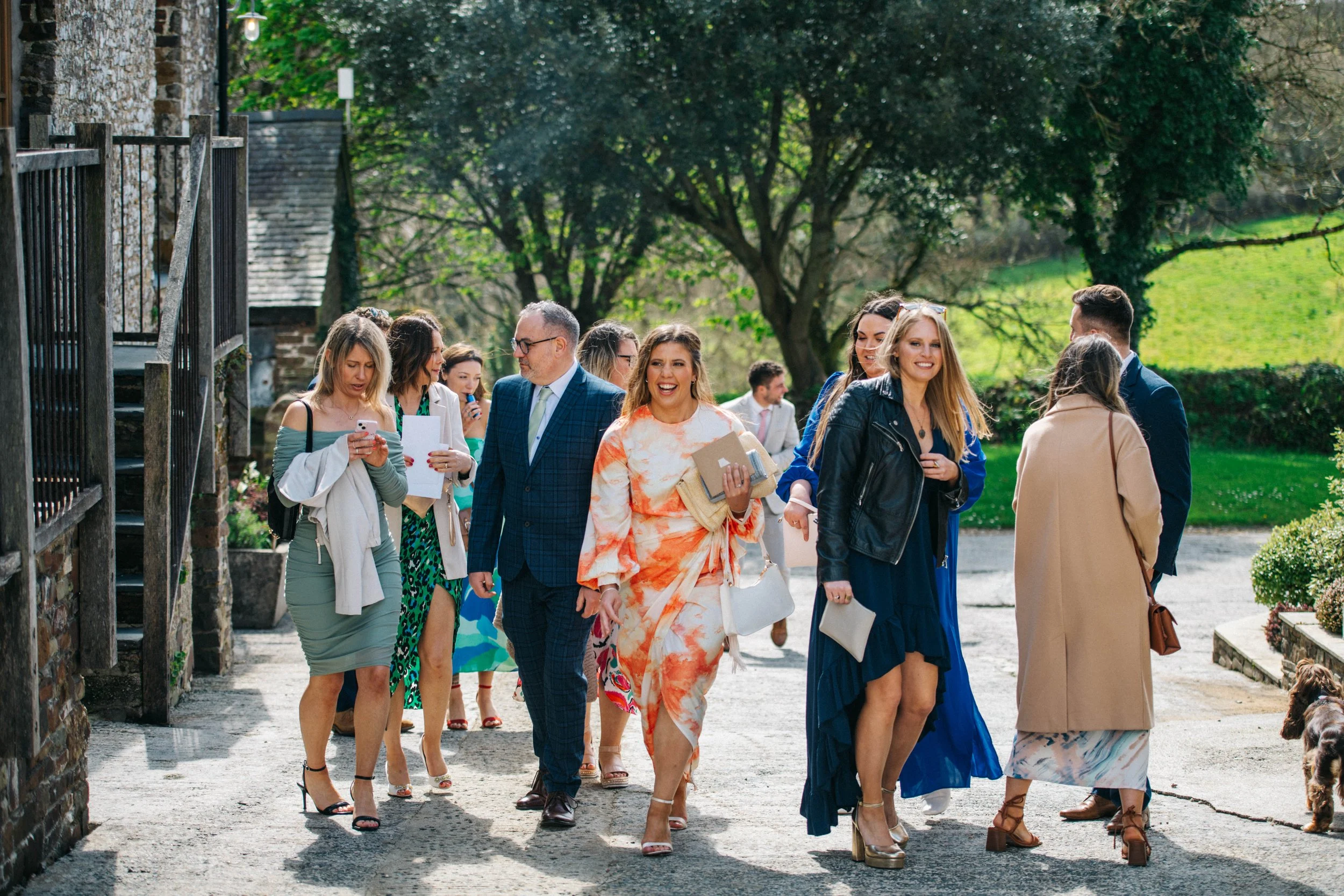 Group of people walking outdoors on a sunny day, dressed in semi-formal and casual attire, with trees and grass in the background.