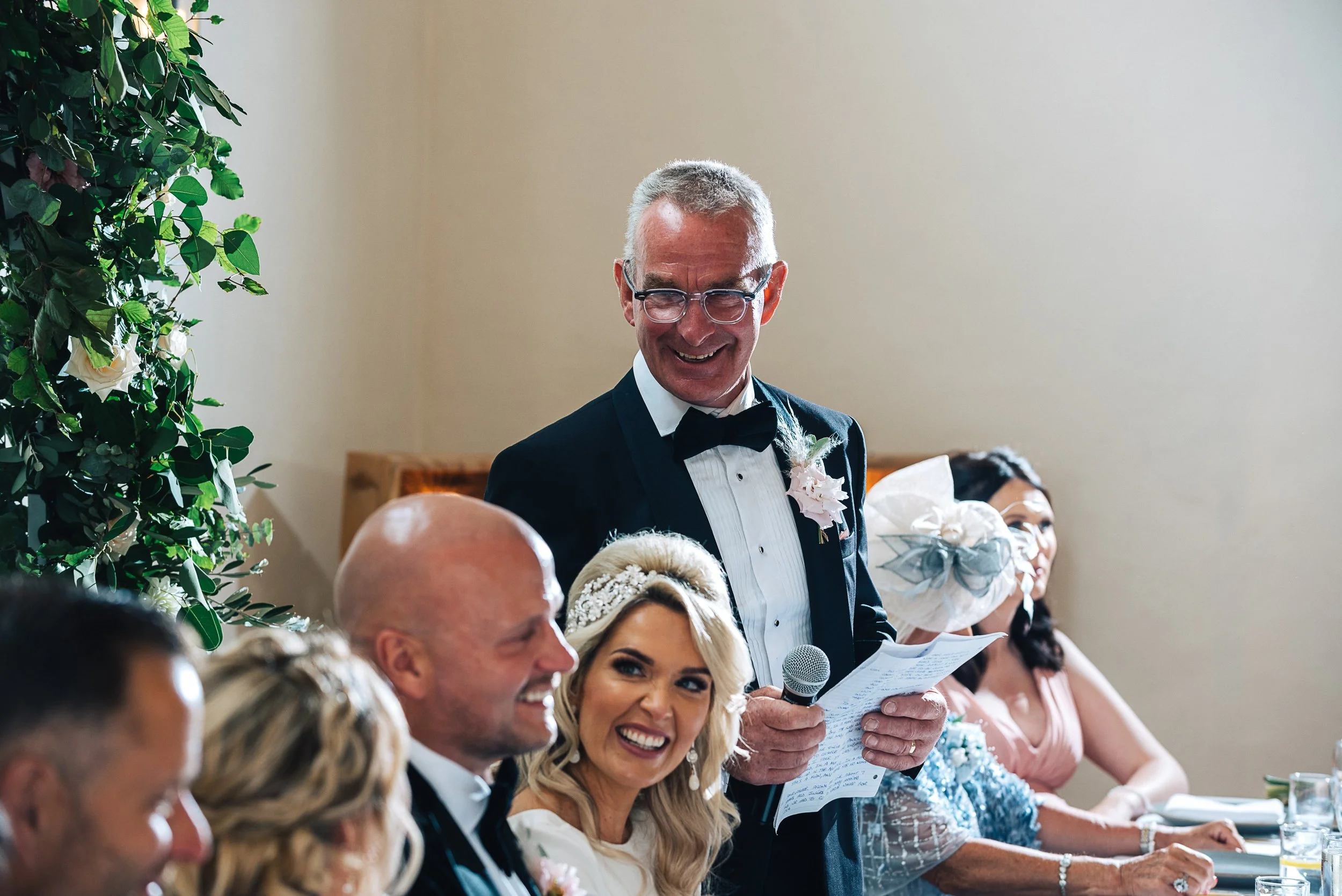A man in a tuxedo giving a speech at a wedding reception, standing next to the bride and groom sitting at a dinner table, with guests around.