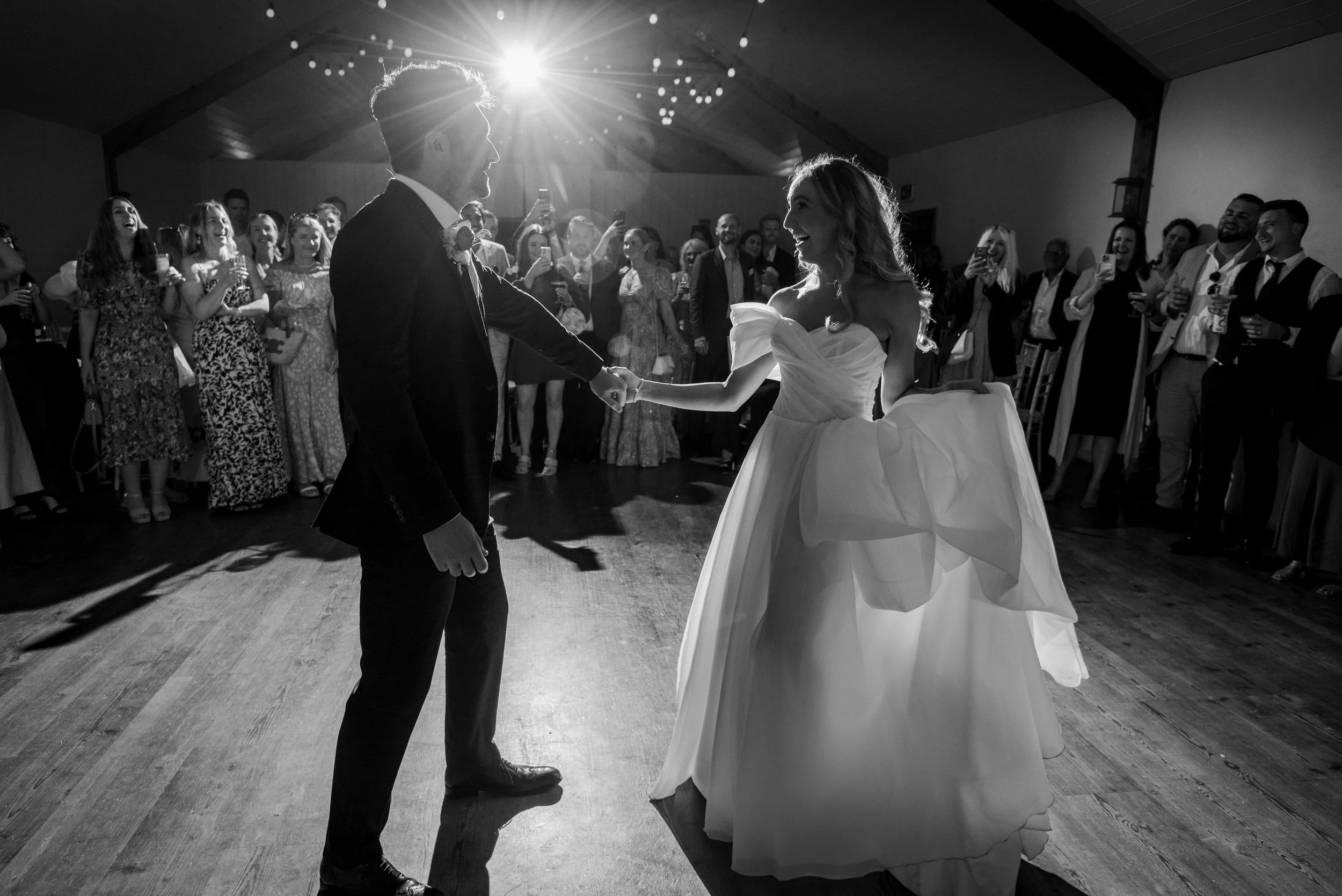 Black and white photo of a bride and groom dancing at their wedding, surrounded by guests, with sunlight shining through the window