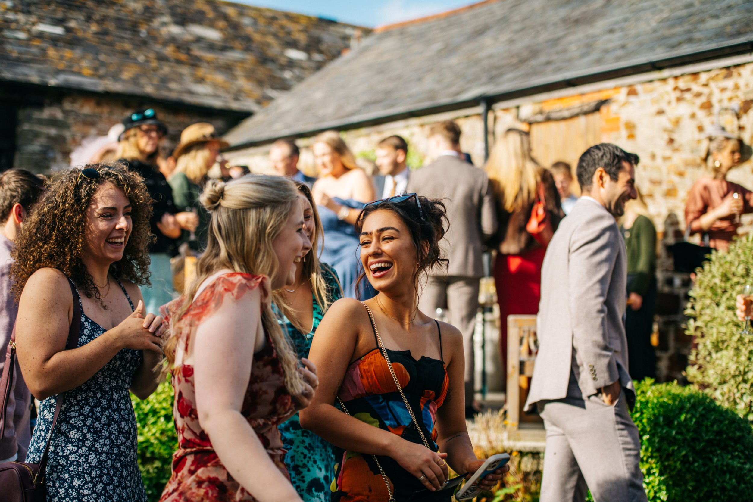 Group of people at an outdoor event, some smiling and laughing, with women in colorful dresses, a man in a gray suit, and rustic buildings in the background.