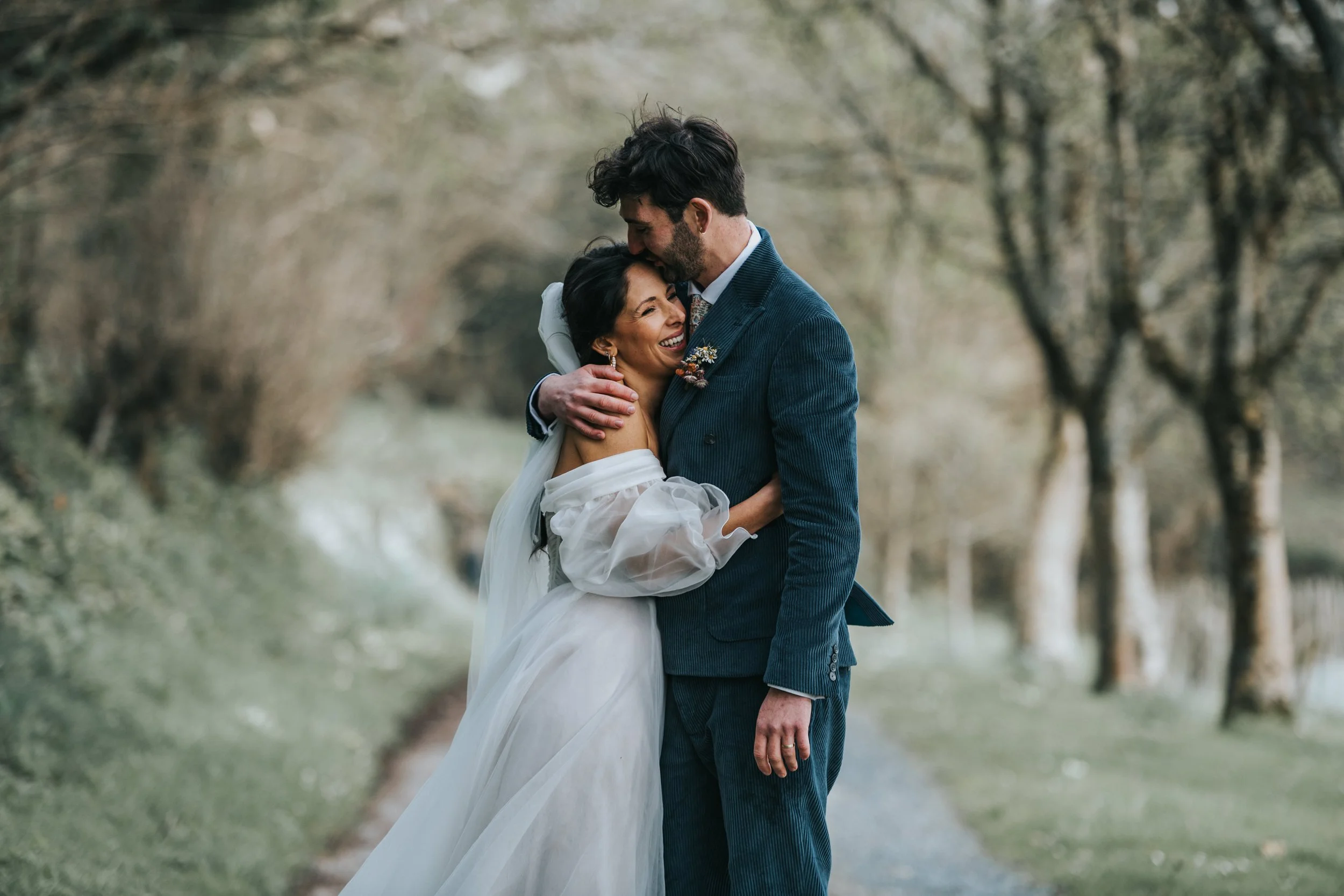A happy bride and groom hugging outdoors on their wedding day, surrounded by trees.