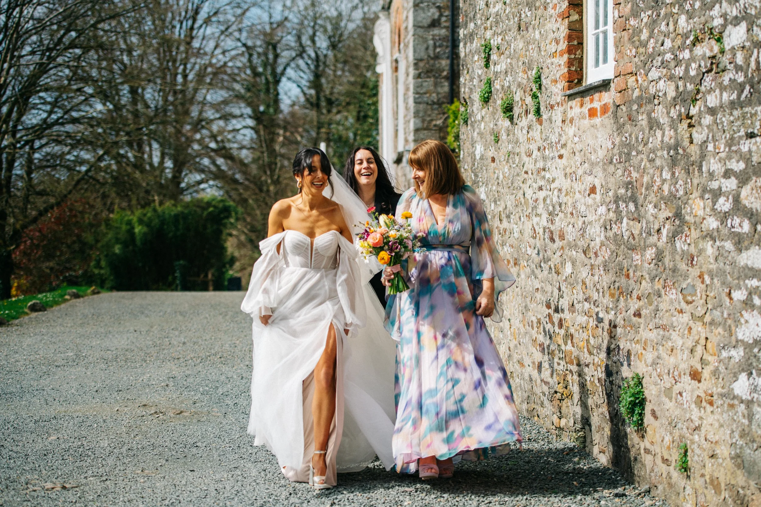 Three women walking outside, one in a wedding dress, holding a bouquet, smiling, with two others smiling and walking with her, beside a stone wall and trees in the background.