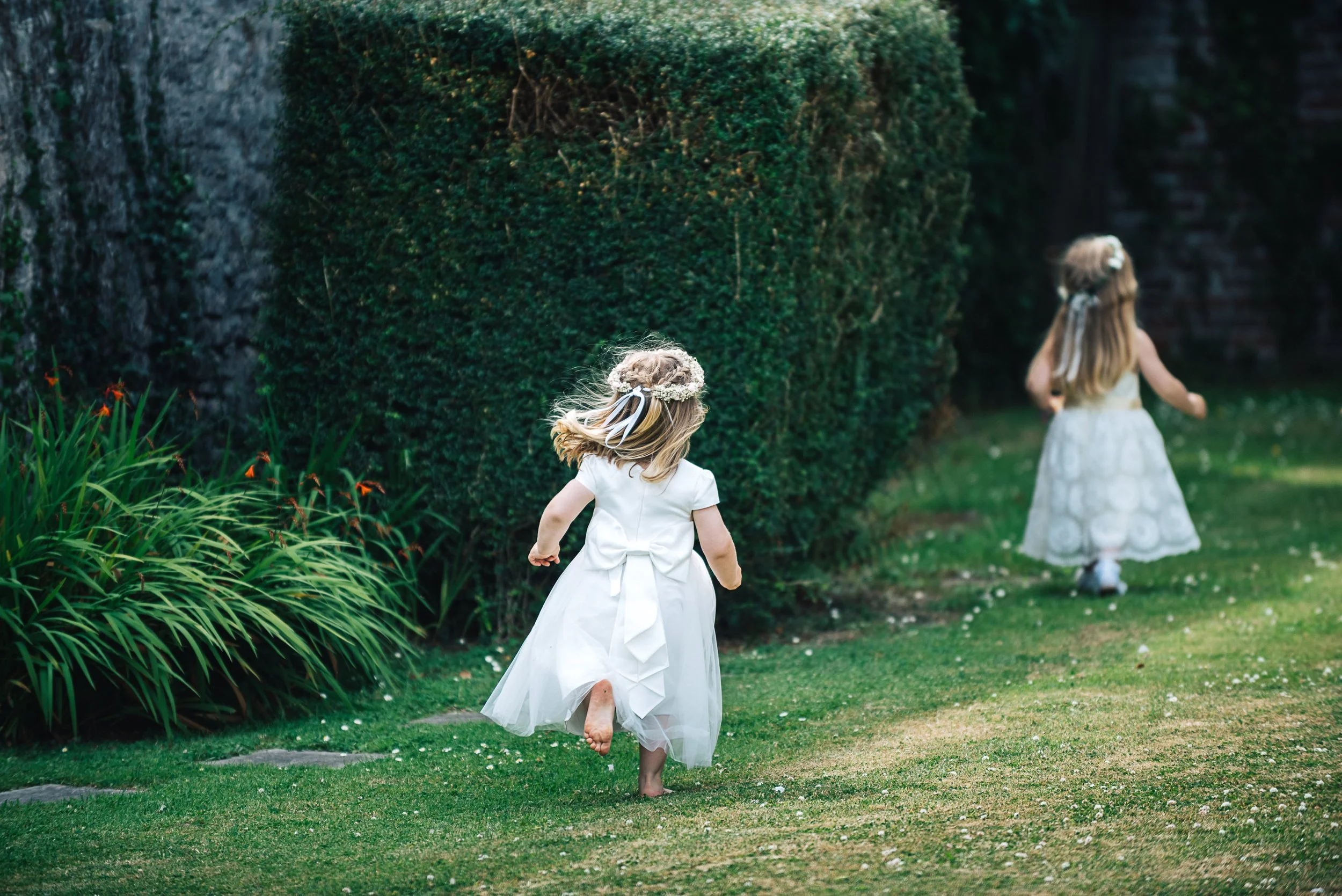 Two young girls in white dresses running on a garden lawn with greenery and hedge in background.