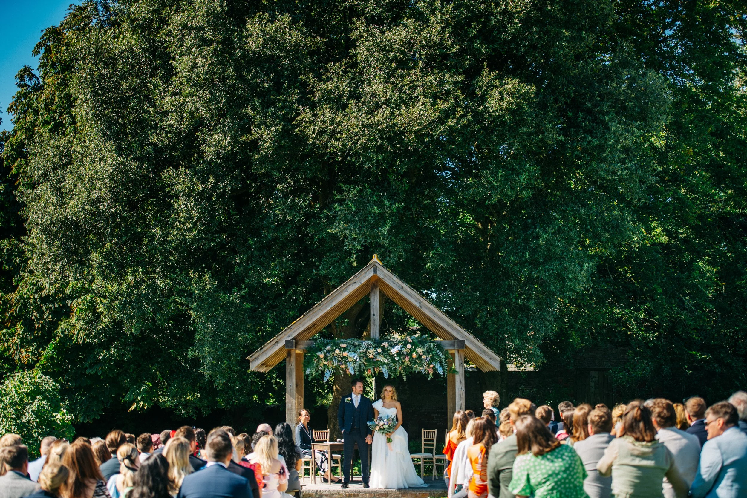 Outdoor wedding ceremony with a bride and groom standing under a wooden arch decorated with flowers, surrounded by seated guests on a sunny day with large green trees in the background.