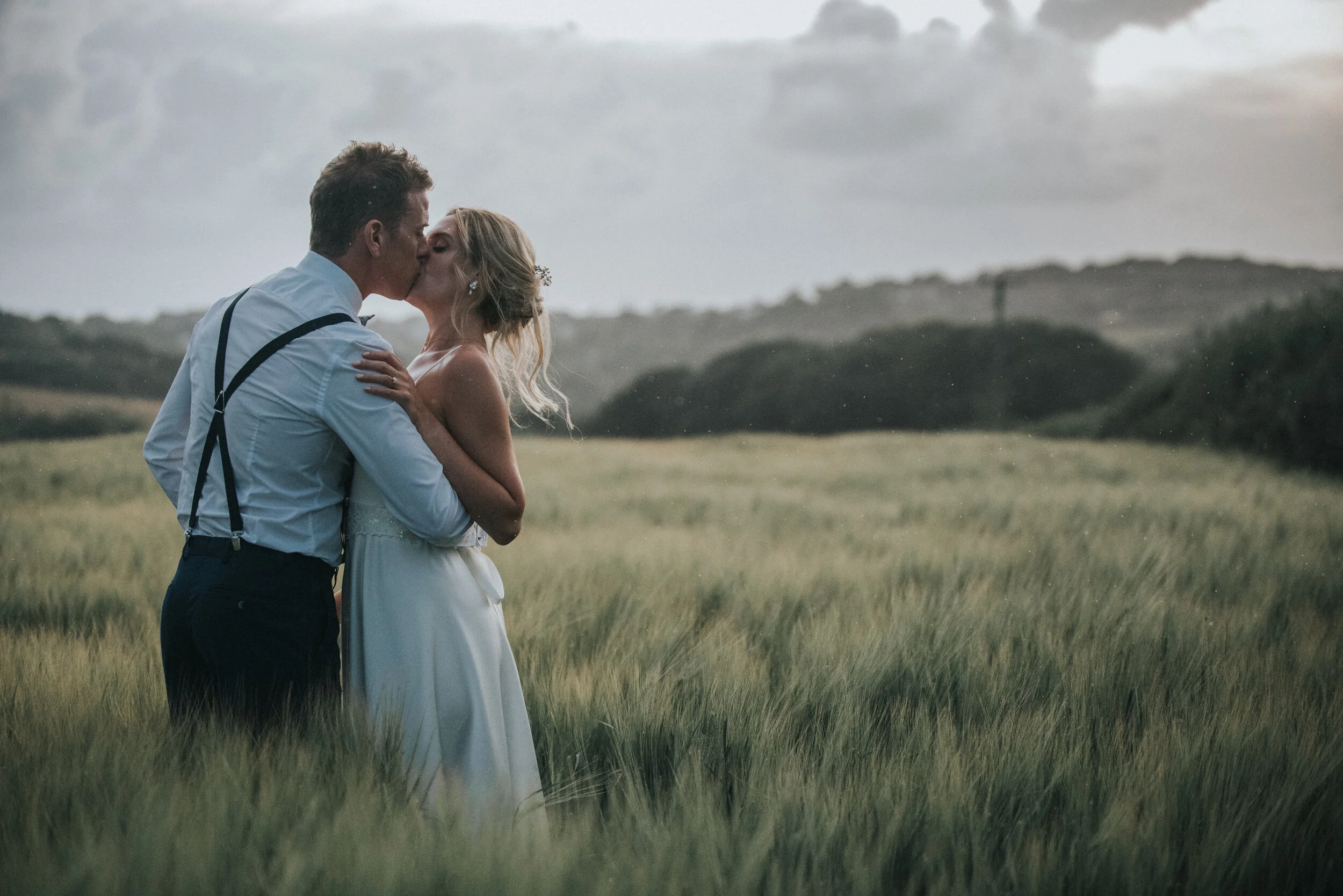 A couple in wedding attire sharing a kiss in a field during overcast weather.