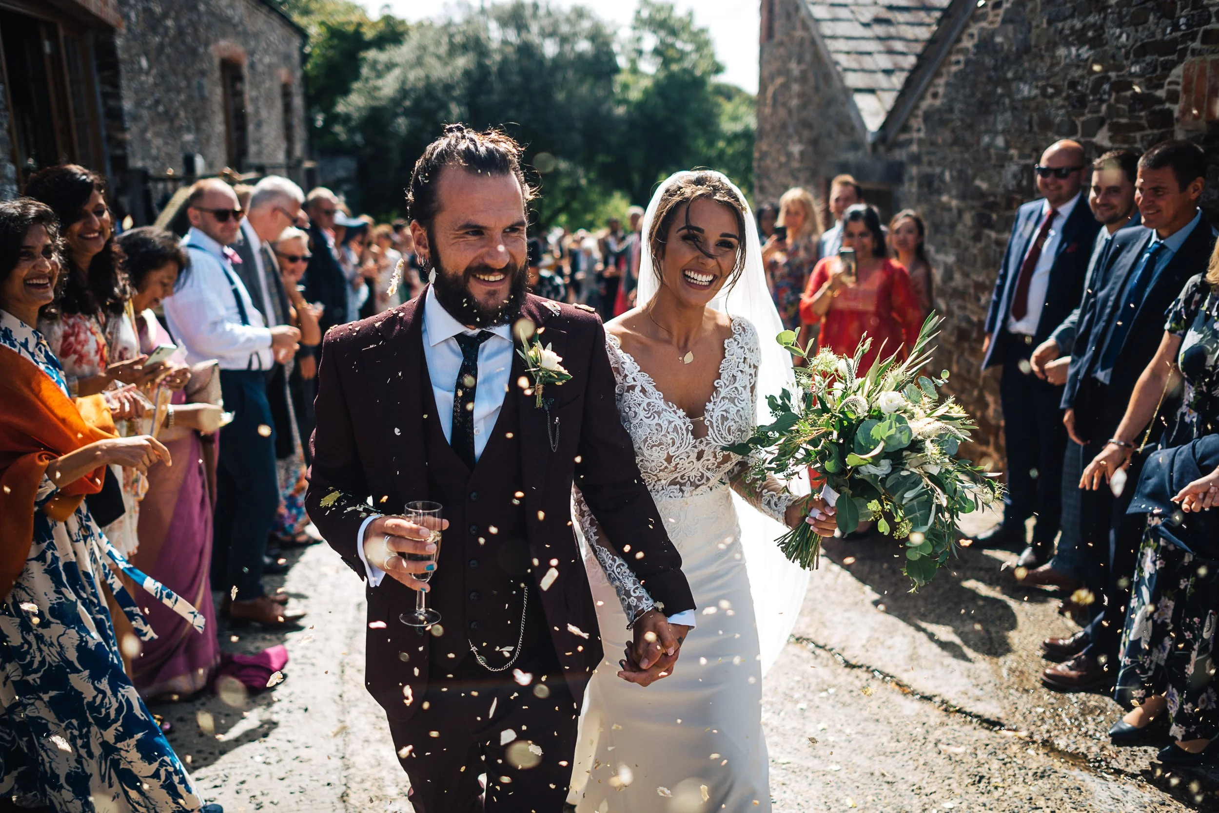 A newlywed couple walks smiling, holding hands, surrounded by guests throwing confetti outdoors on a sunny day.