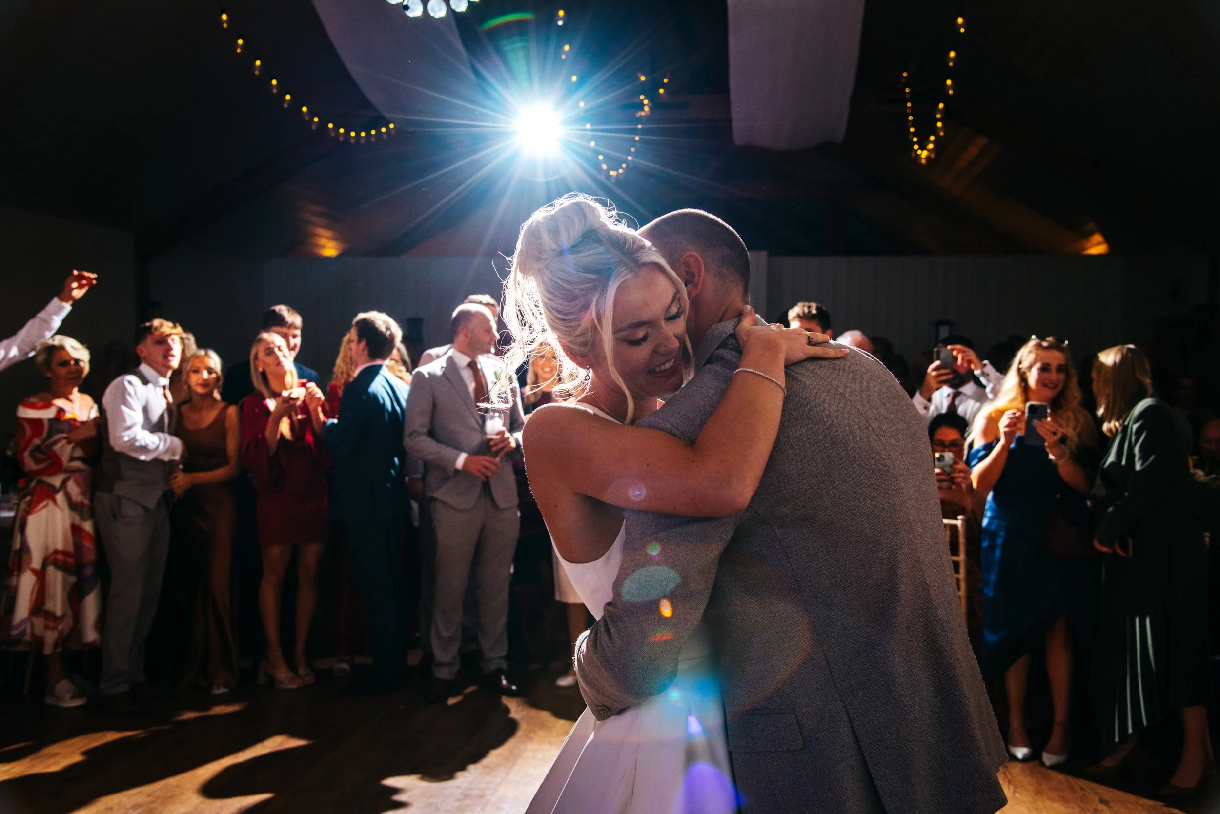 A bride and groom are dancing closely at their wedding reception, surrounded by friends and family who are watching and taking photos. A bright light shines from above, creating a romantic atmosphere.