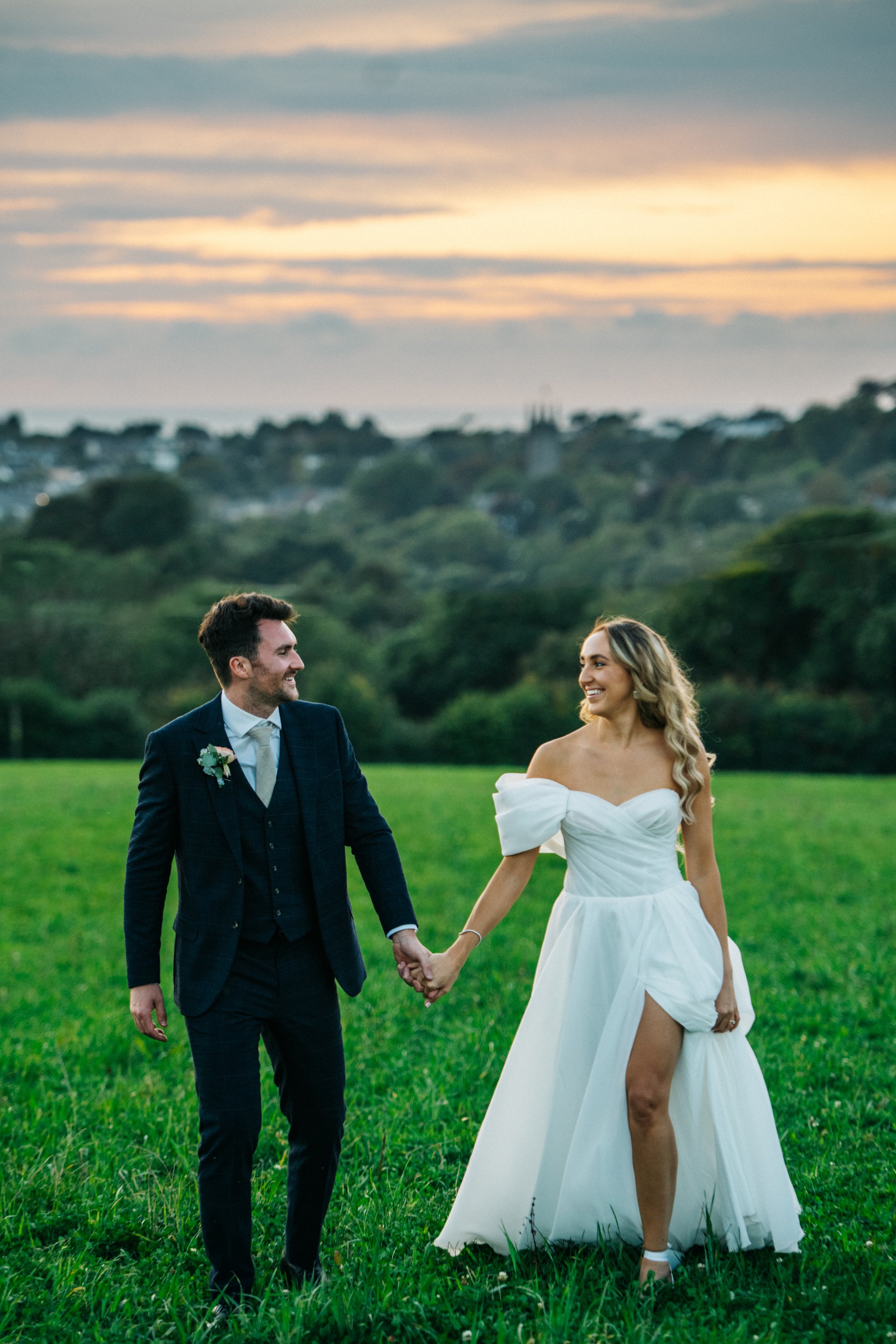 A bride and groom holding hands and walking in a grassy field at sunset.