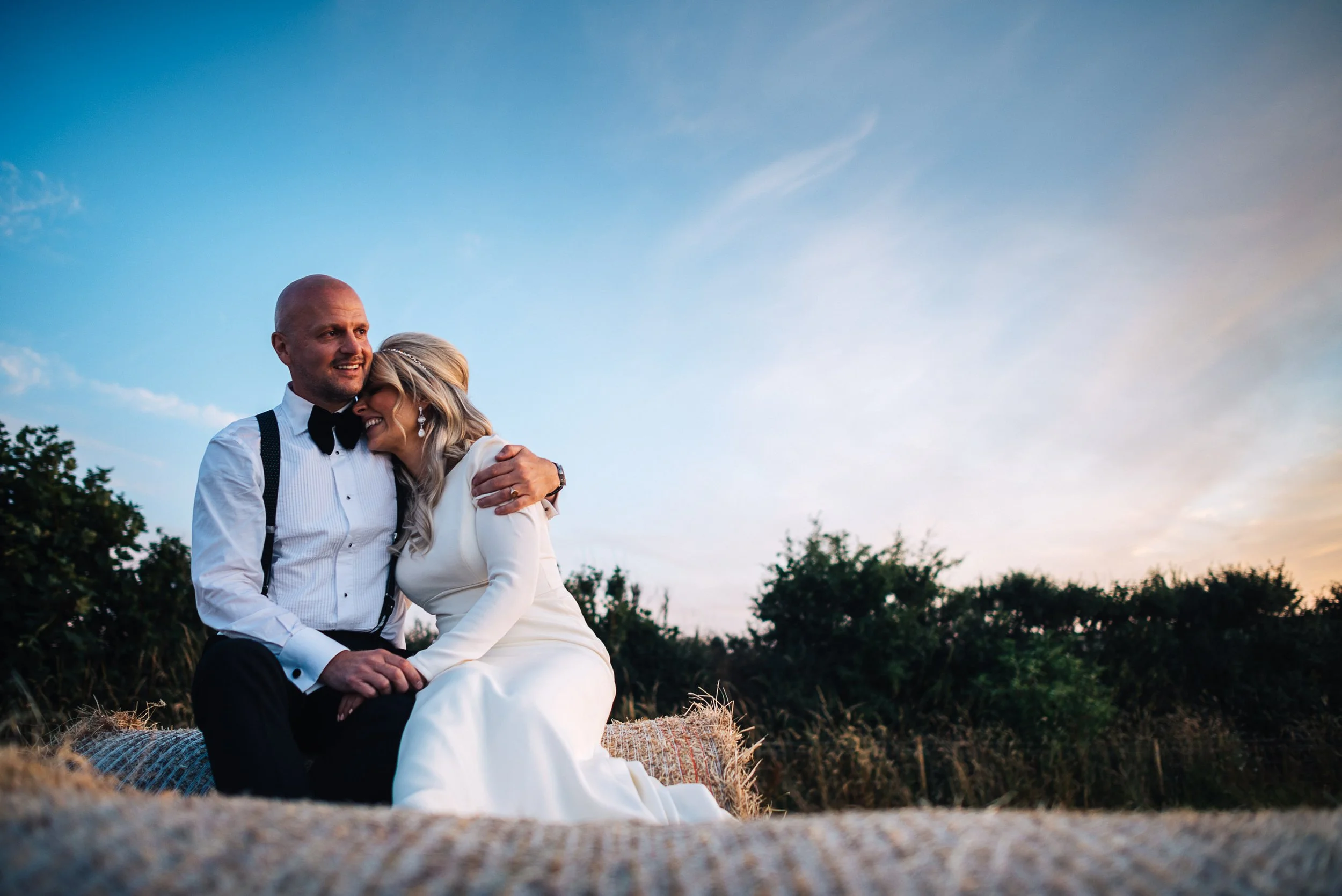 a couple sitting on a hay bale outdoors during sunset, embracing each other, the woman is wearing a white dress and the man is wearing a white shirt with black suspenders and a bow tie